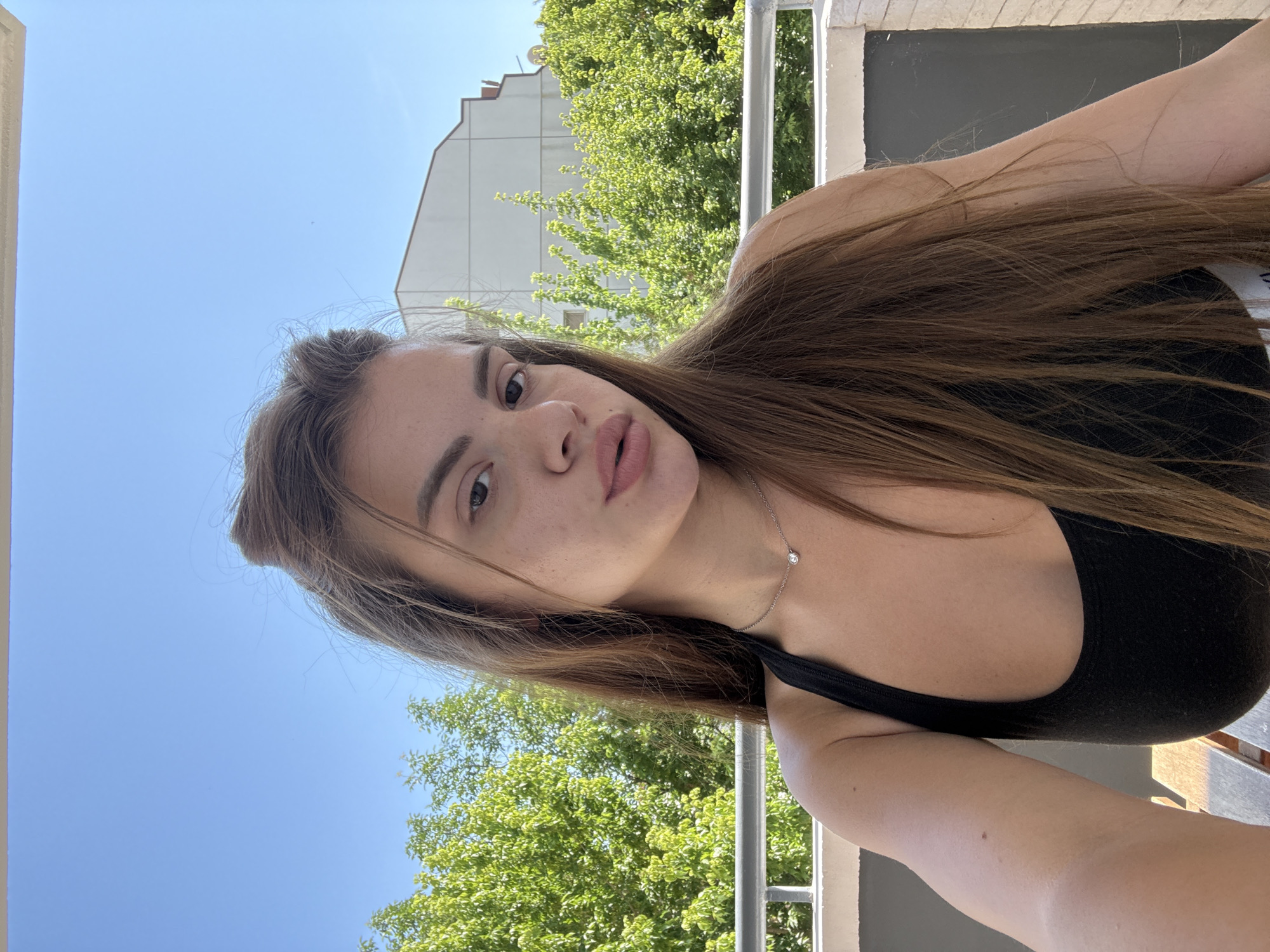 A young woman with long brown hair, wearing a black tank top and a delicate necklace with a single pendant, is taking a selfie on a balcony. She is looking directly at the camera with a neutral expression. Behind her, lush green trees and a white building are visible under a clear blue sky. The scene suggests a sunny day, possibly in an urban setting, given the building in the background.