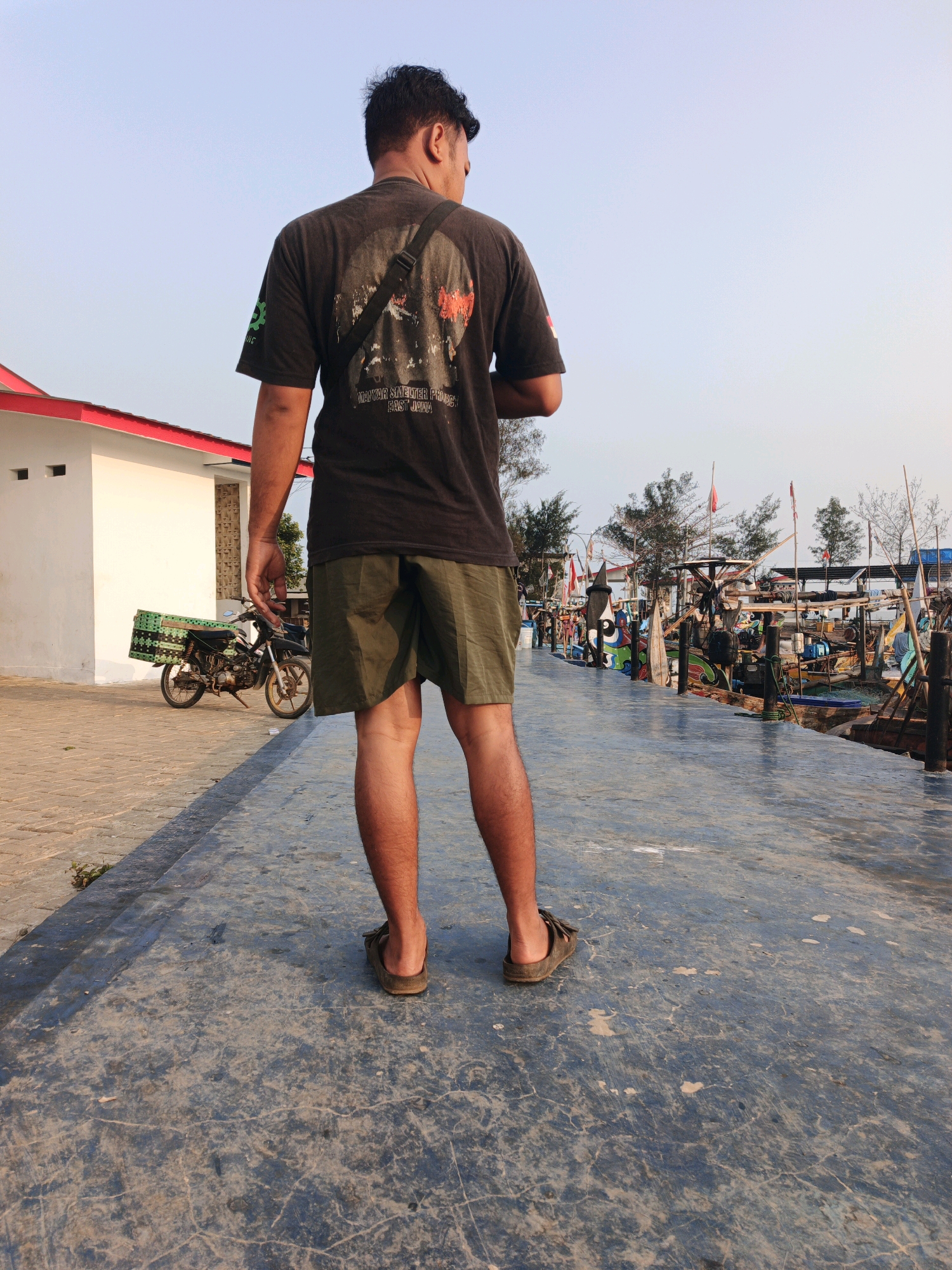 A man stands facing away from the camera on a paved walkway. He is wearing a dark t-shirt with a graphic on the back, olive green shorts, and brown sandals. A black strap is slung across his chest. To his left, a white building with a red trim and a motorcycle are visible. The motorcycle has a green crate attached to the back. To his right, a harbor filled with numerous fishing boats is situated along the walkway. The sky is clear and light blue. The scene appears to be in a coastal area of Indonesia.