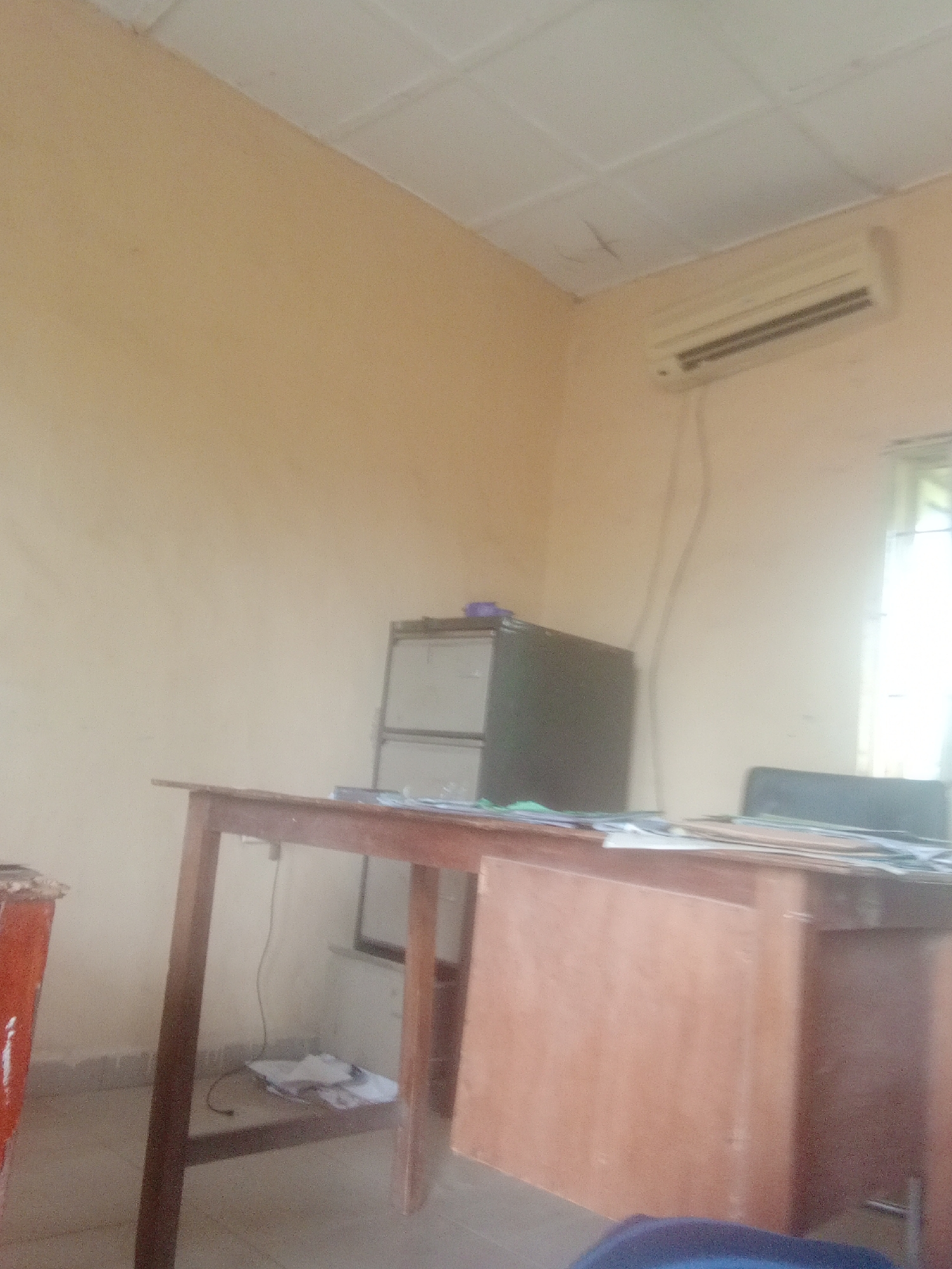 The image depicts a room, likely an office or study, with plain, light-colored walls and a drop ceiling. On the wall above a wooden desk is an air conditioning unit. The desk itself is a long, simple wooden structure with a wooden panel on one side, likely part of a larger desk setup. Papers and some folders are stacked on the desk. To the left of the desk, a filing cabinet with multiple drawers is visible. A large, orange cylindrical object, possibly a waste bin or a decorative item, is partially visible on the far left. In the background, a window with diffused light suggests it could be daytime, though the interior lighting appears to be the primary illumination. The overall atmosphere is functional and somewhat sparse. There is no visible text in the image. The context suggests this scene is in Jalingo, Nigeria.