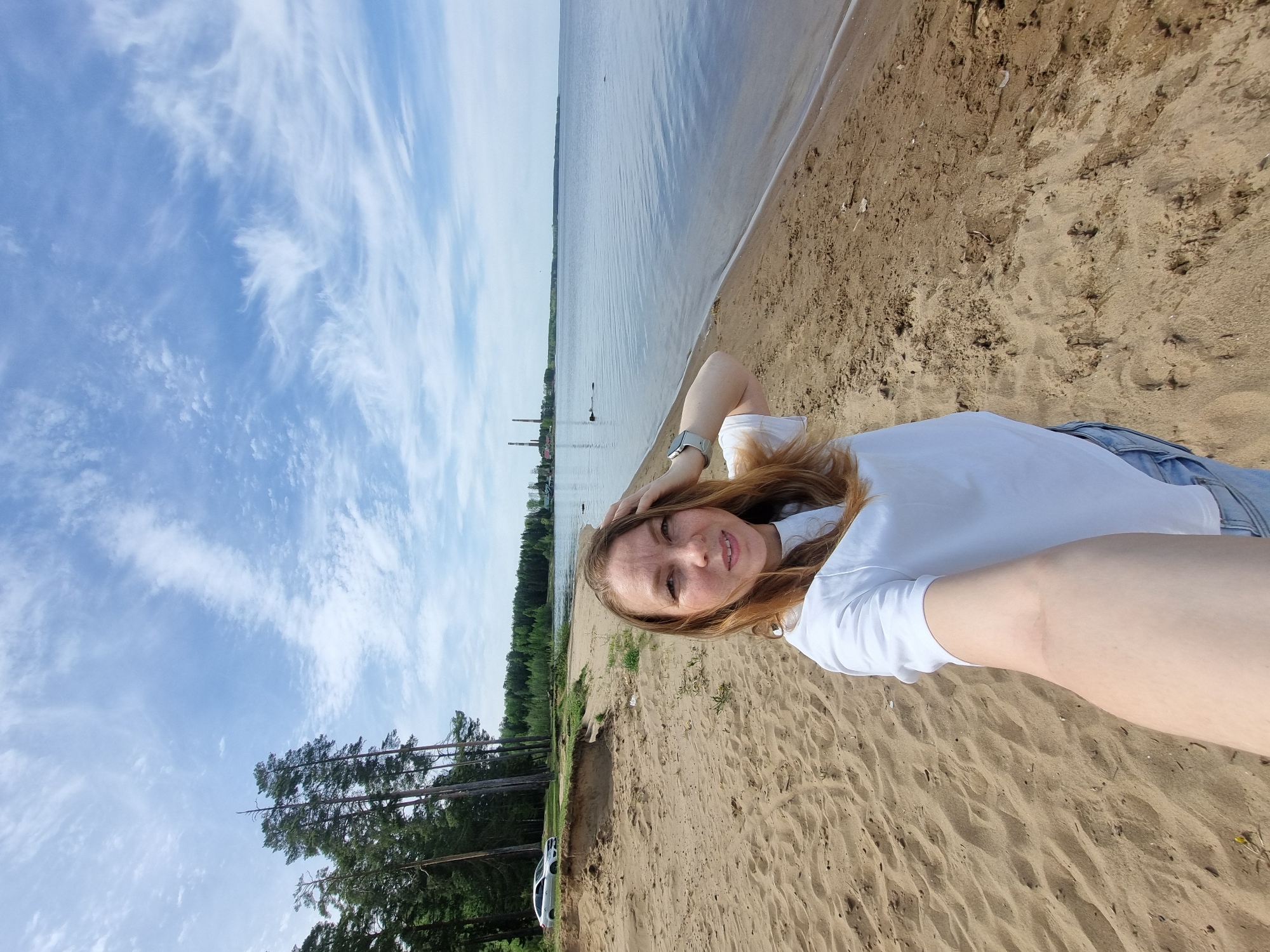 A woman lies on a sandy beach. The location is likely in the city of Chaikovsky, Russia, based on visual cues (landscape, architecture) within the wider context of the original query. The woman is wearing a white T-shirt and light-colored pants. Her auburn hair is spread on the sand. She holds her head with her hand. The background includes a body of water, possibly a river or lake, with forest visible on the opposite shore. The sky is blue with scattered clouds. To the left of the frame, a white car is parked near trees. Industrial structures (potentially factory chimneys) are vaguely visible in the distance across the water.