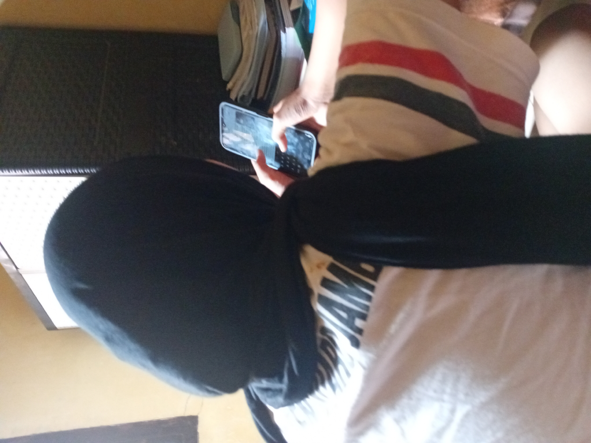 A person wearing a black hijab and a white t-shirt with black and red stripes is sitting and looking at a smartphone. The t-shirt has black lettering that appears to say "JAMB". The background includes a dark woven cabinet and a stack of books or papers on a surface. The lighting suggests an indoor setting. This scene is likely located in Kasang Pudak, Indonesia.