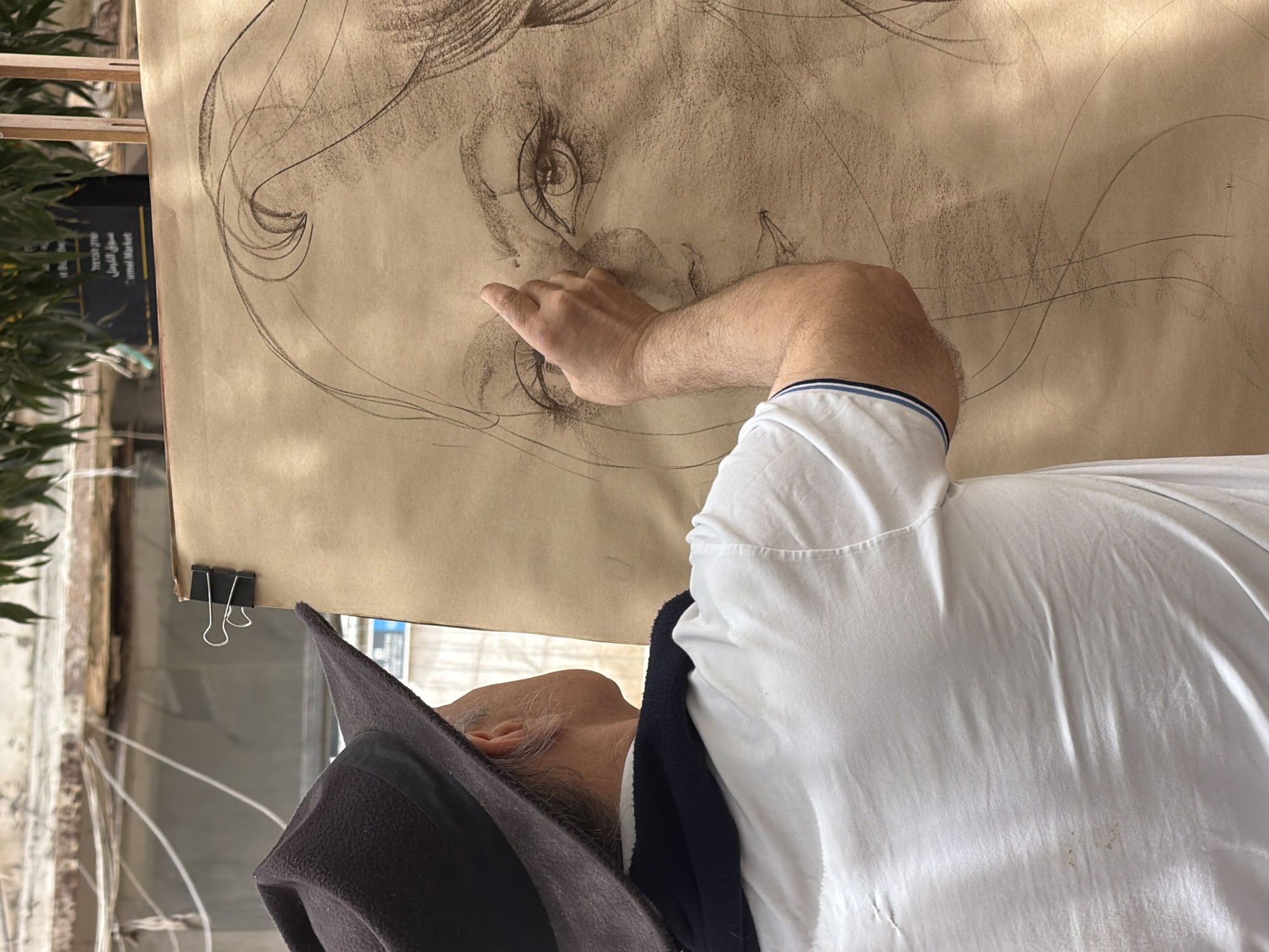 An artist wearing a black hat and white shirt is standing in front of a large paper affixed to a stand. The paper has a charcoal drawing of a woman's face. The artist is using his fingers to smudge and blend the charcoal. The location appears to be an outdoor setting in Tel-Aviv, Israel, indicated by the Hebrew signage visible in the background. The scene is likely a street art display or a live drawing session.