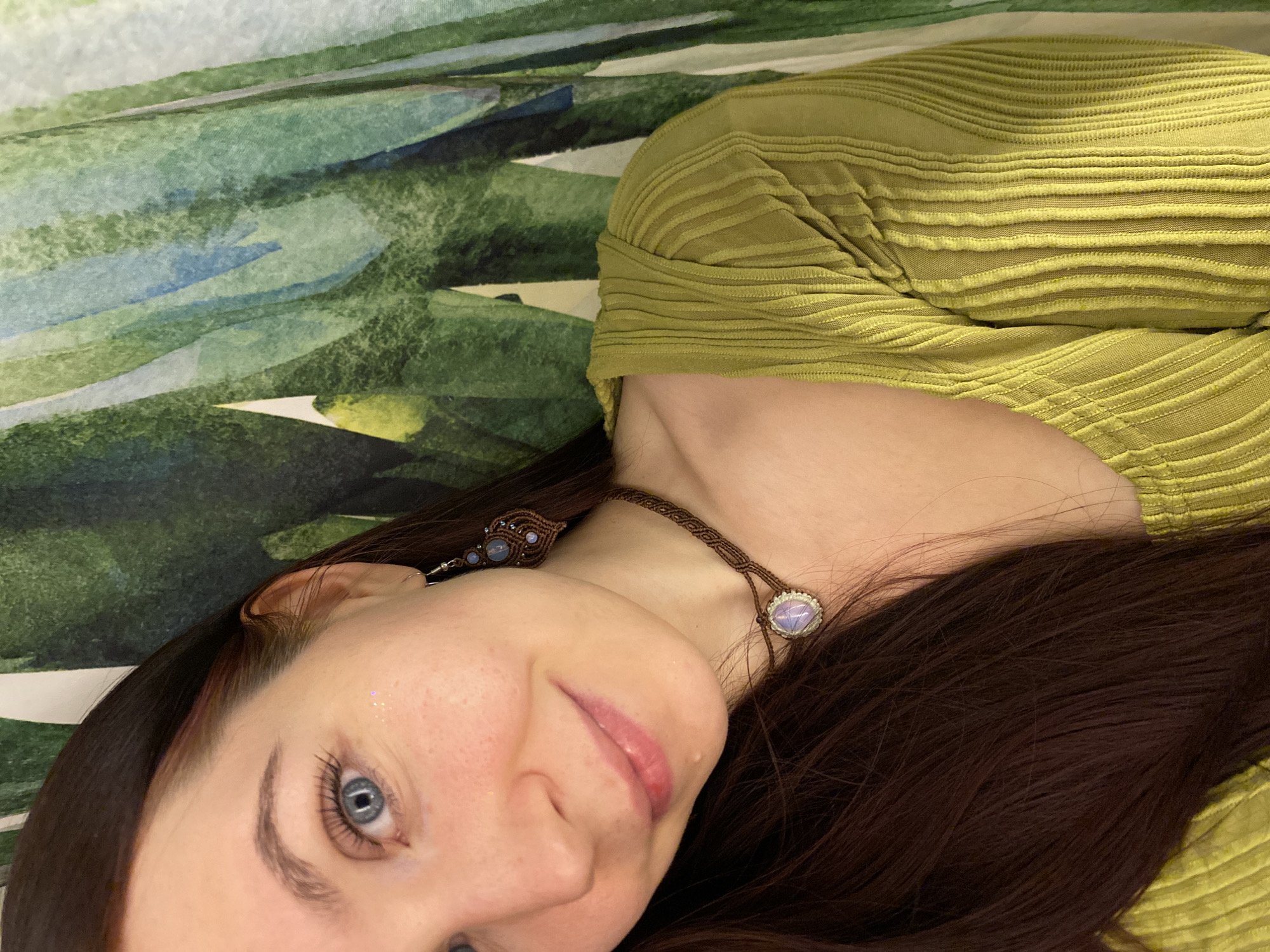 A woman is depicted from a close-up perspective, turned slightly towards the right. She has fair skin, blue eyes, and auburn hair. There are faint sparkles on her cheek. She is wearing a green, textured top and a macrame necklace with a pale gemstone. The background consists of a blurred, patterned fabric featuring green, blue, and white hues. The location is not discernible from the image, nor can it be determined whether it is in Chaikovsky, Russia.