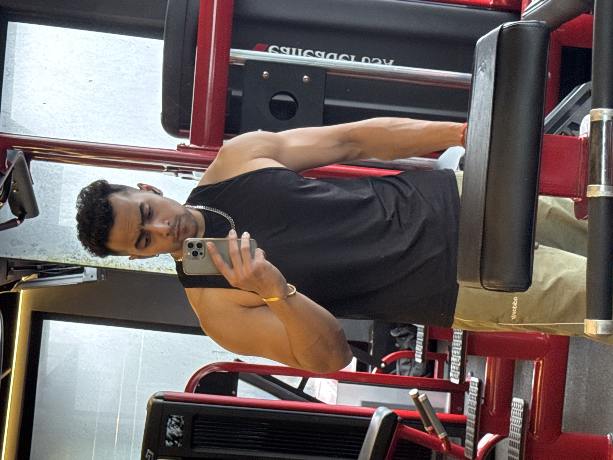 A man is taking a mirror selfie in a gym. He is wearing a black sleeveless t-shirt, tan athletic shorts, and a silver chain necklace. He has curly black hair and is holding a silver smartphone in his right hand, which is raised to take the photo. His left arm is bent at the elbow, with his hand resting on his leg. He is wearing a gold bracelet on his left wrist. The gym equipment around him is red and black. The flooring appears to be grey. The lighting is bright and appears to be from overhead fixtures. The background shows workout machines and a large reflective surface, likely a mirror or window. There are no other people visible in the image. The overall scene depicts a person in a fitness environment.