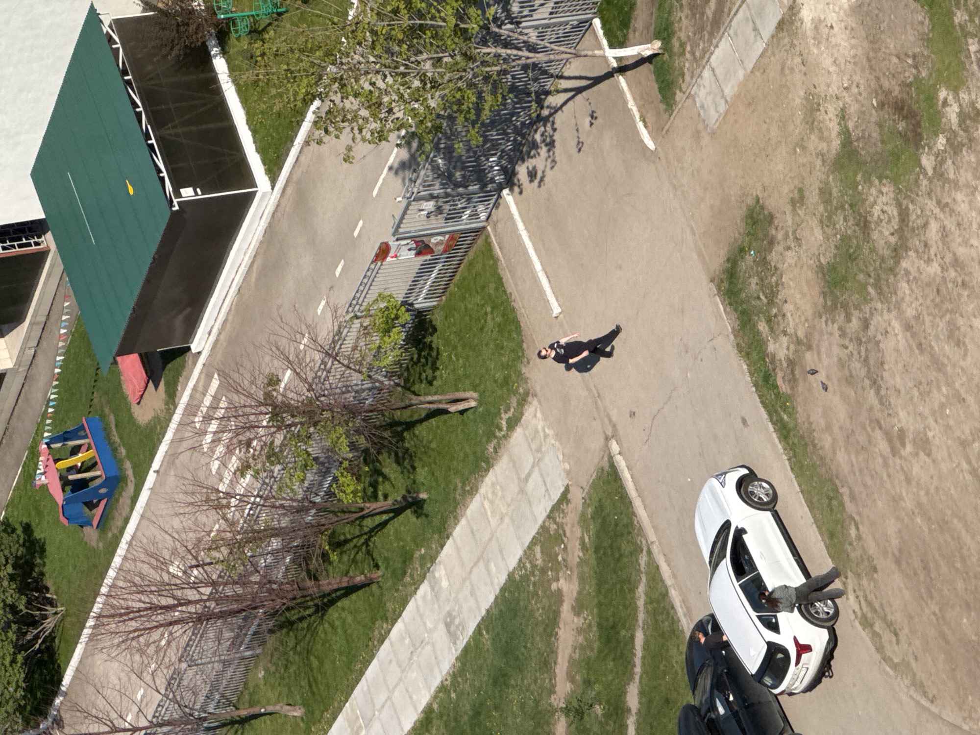 An aerial shot depicts a mixed urban landscape, with paved pathways intersecting green spaces. A solitary person in dark clothing is seen walking along one of the paths. Adjacent to the path, a white car is parked, and nearby is a dark-colored vehicle. A fenced playground area with colorful equipment is visible on the left side of the image. There is also a large green-roofed structure. Trees with budding leaves dot the landscape, and the overall impression is one of a park or recreational area within a city.