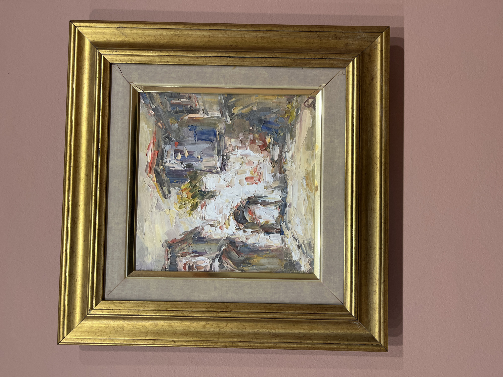 An impressionistic oil painting, framed in ornate gold, hangs on a solid dusty rose-colored wall. The artwork, characterized by thick impasto brushstrokes, depicts what appears to be an outdoor bustling scene, possibly a street, market, or cafe. Dominant colors within the painting include whites, creams, blues, grays, and muted yellows, with accents of red and green. The abstract nature of the brushwork creates a sense of movement and light, hinting at figures, tables, or architectural elements without explicit detail.
The painting is housed within a substantial gilded frame featuring multiple ridges and a slightly aged or distressed finish. Inside the gold frame is a lighter, neutral-toned linen mat, providing a subtle transition to the artwork. One of the cropped images reveals a small crack or imperfection in the upper left miter joint of the inner frame. The overall presentation is classic and well-maintained, suggesting an indoor setting for display. No text is visible on the painting, frame, or wall. The setting for this media file is Plovdiv, Bulgaria.