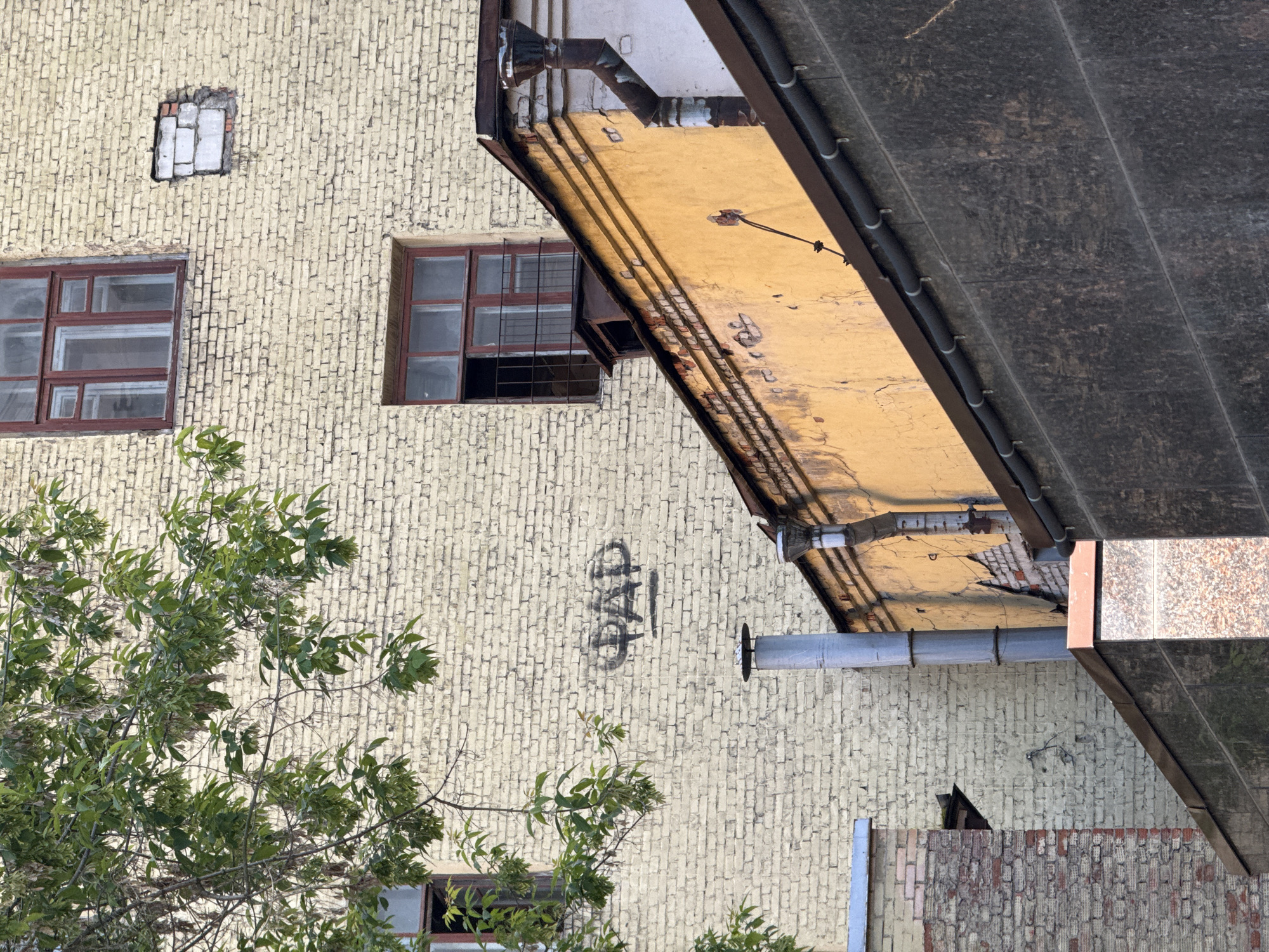 Here is a forensic description of the image:
The image is a medium shot of the exterior of a building, potentially located in Moscow, Russia. The building's facade is predominantly pale yellow brick, showing signs of wear and age. There are several windows visible. Two are intact with brown frames, and one is partially bricked over. Black graffiti is present on the brick wall.
A structure overhangs the wall, painted yellow on the underside. Attached to the underside are a metal pipe and other mechanical elements. A dark, sloped awning is positioned above the overhang, further obstructing the view.
In the foreground, the branches and foliage of a tree are visible, partially obscuring the building. Another brick wall, painted red and yellow is visible at the bottom of the image.