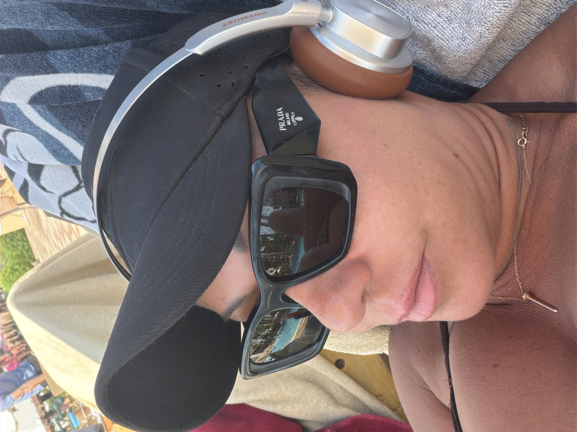 A close-up, eye-level, outdoor, sunny shot shows a person lying down, wearing a black baseball cap, large black sunglasses with "PRADA MILANO" written on the side, and silver and brown headphones. The person's face is partially visible, with closed eyes, and they are wearing a gold necklace with a pointed pendant. The background shows a swimming pool with blue water and some palm trees. The person appears to be relaxing on a lounge chair, possibly at a resort in Sharm El Sheikh, Egypt.