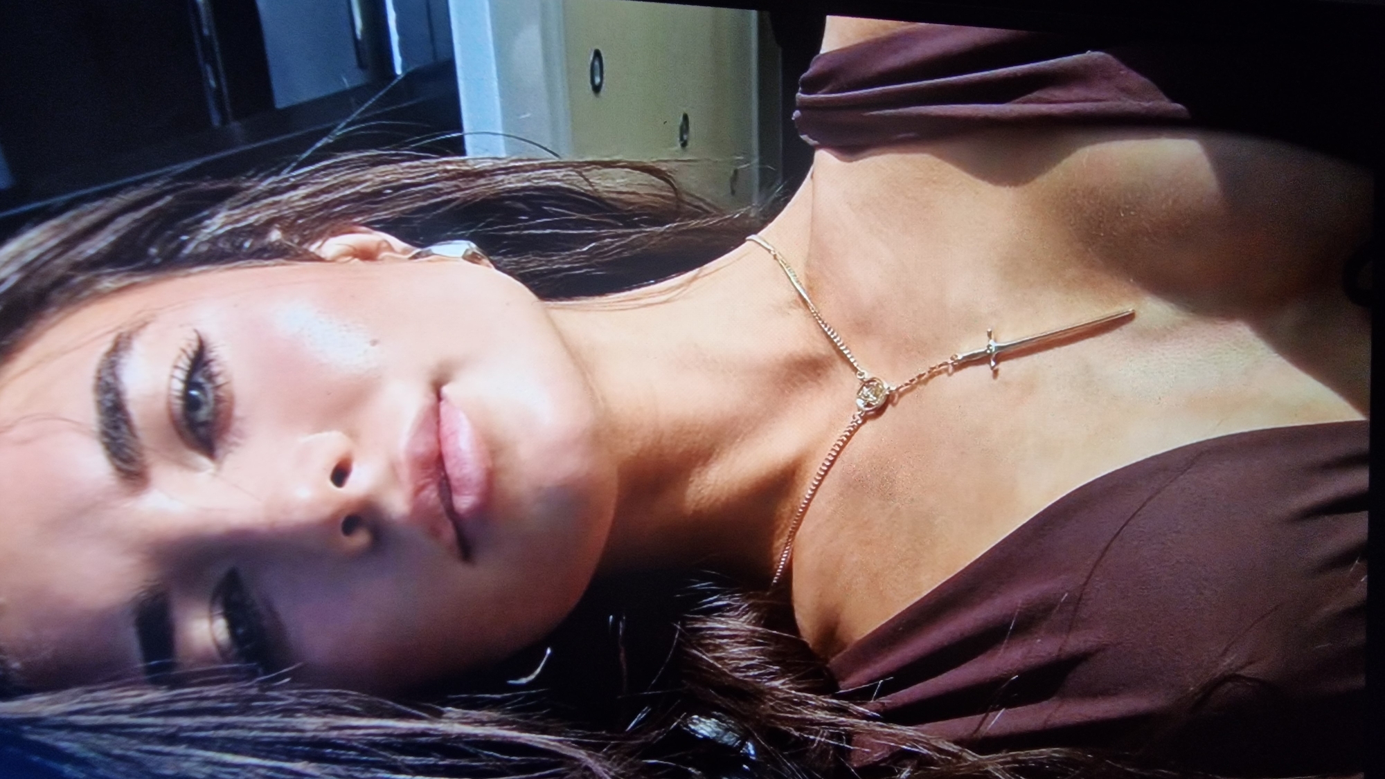 This is a close-up, head-and-shoulders shot of a young woman with dark hair and blue-green eyes. She is wearing a dark brown, possibly silk, top and a gold necklace with a pendant shaped like a sword. She has a subtle makeup application with eyeliner and mascara. Her skin appears dewy and highlighted. The background is blurred and appears to be outdoors, possibly on a boat or near water, with hints of dark structures and bright reflections. No specific location in Aksu City, China, can be determined from the image.