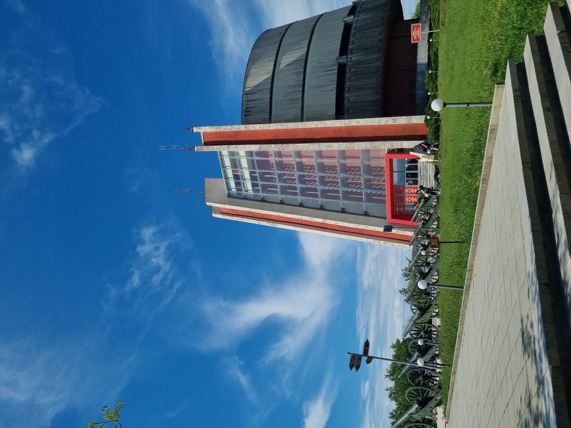 The image shows the Pleven Panorama, a large monument in Pleven, Bulgaria. The monument consists of a large concrete cylinder with a glass-fronted building attached to its side. In front of the monument are a series of cannons lined up along a paved walkway. The background is a bright blue sky with scattered clouds. The scene is outdoors and appears to be taken during the day.
