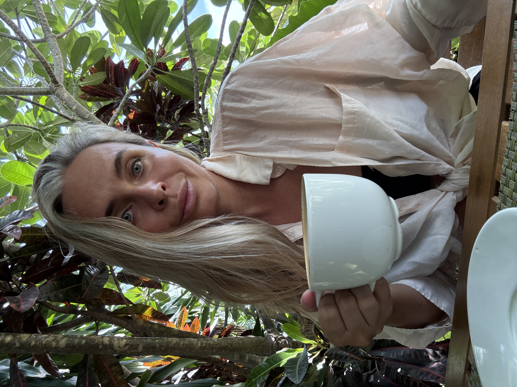 A middle-aged Caucasian woman with long blonde hair and blue eyes is sitting outdoors, holding a white coffee mug in her left hand. She is wearing a light-colored, loose-fitting shirt tied at the waist. The background consists of lush green foliage and tree branches, suggesting a tropical environment, likely in the Ko Samui District of Thailand. Sunlight filters through the leaves, creating dappled light on the scene. A white saucer is visible on the table in front of her.