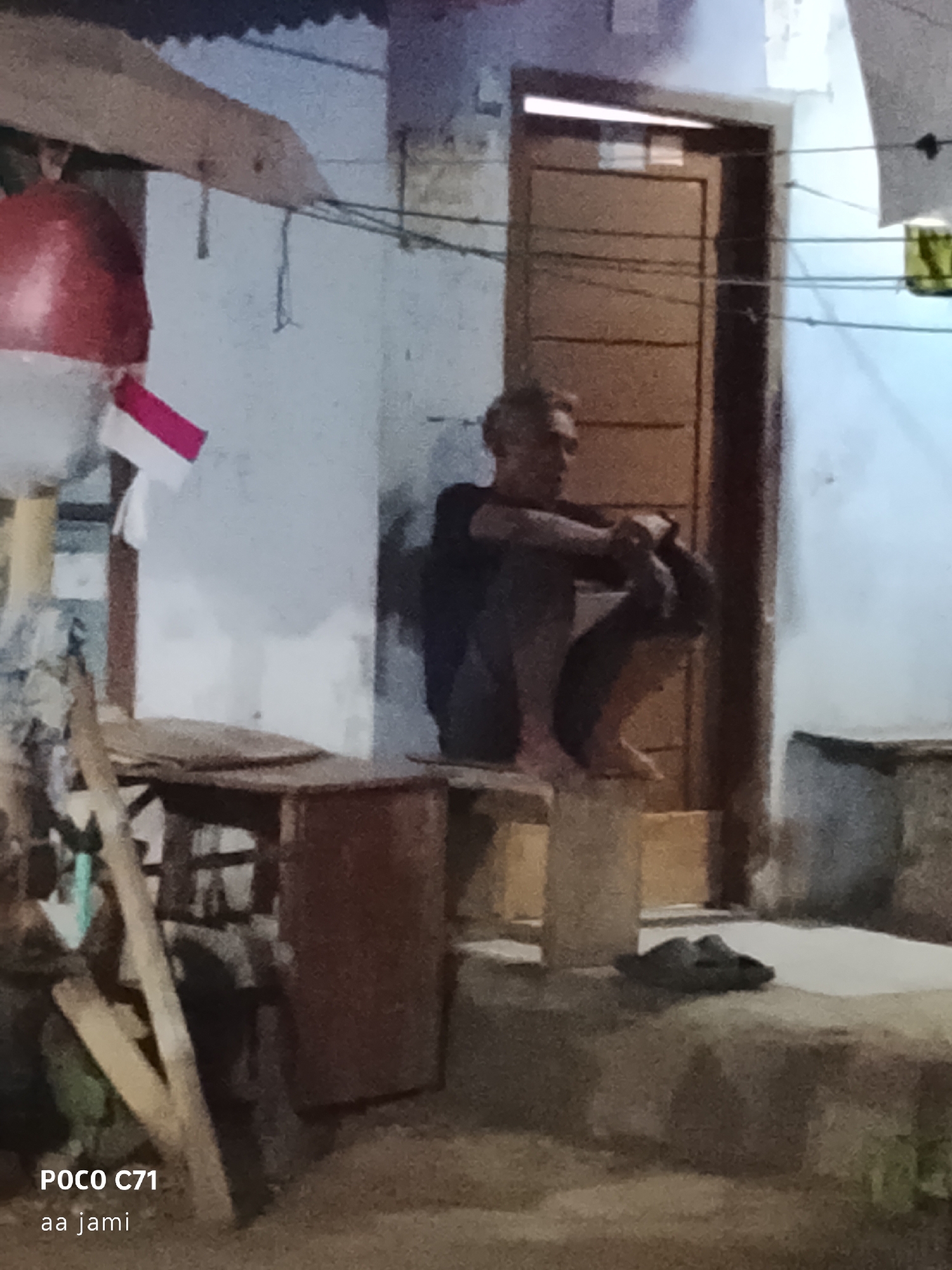 A person is squatting on a wooden structure in front of a building with a brown door. The person is wearing dark clothing and has their arms wrapped around their legs. To the left of the person, there is a red object, possibly a buoy, with a white and red flag attached to it. In the foreground, there are some wooden planks and what appears to be a metal drum. On a raised platform to the right, a pair of dark-colored sandals are visible. The background shows a wall and a door. The text "POCO C71 aa jami" is overlaid on the bottom left corner of the image. The scene appears to be an outdoor area in Tasikmalaya, Indonesia, possibly near a roadside or informal settlement, given the rustic elements and the presence of the Indonesian flag.