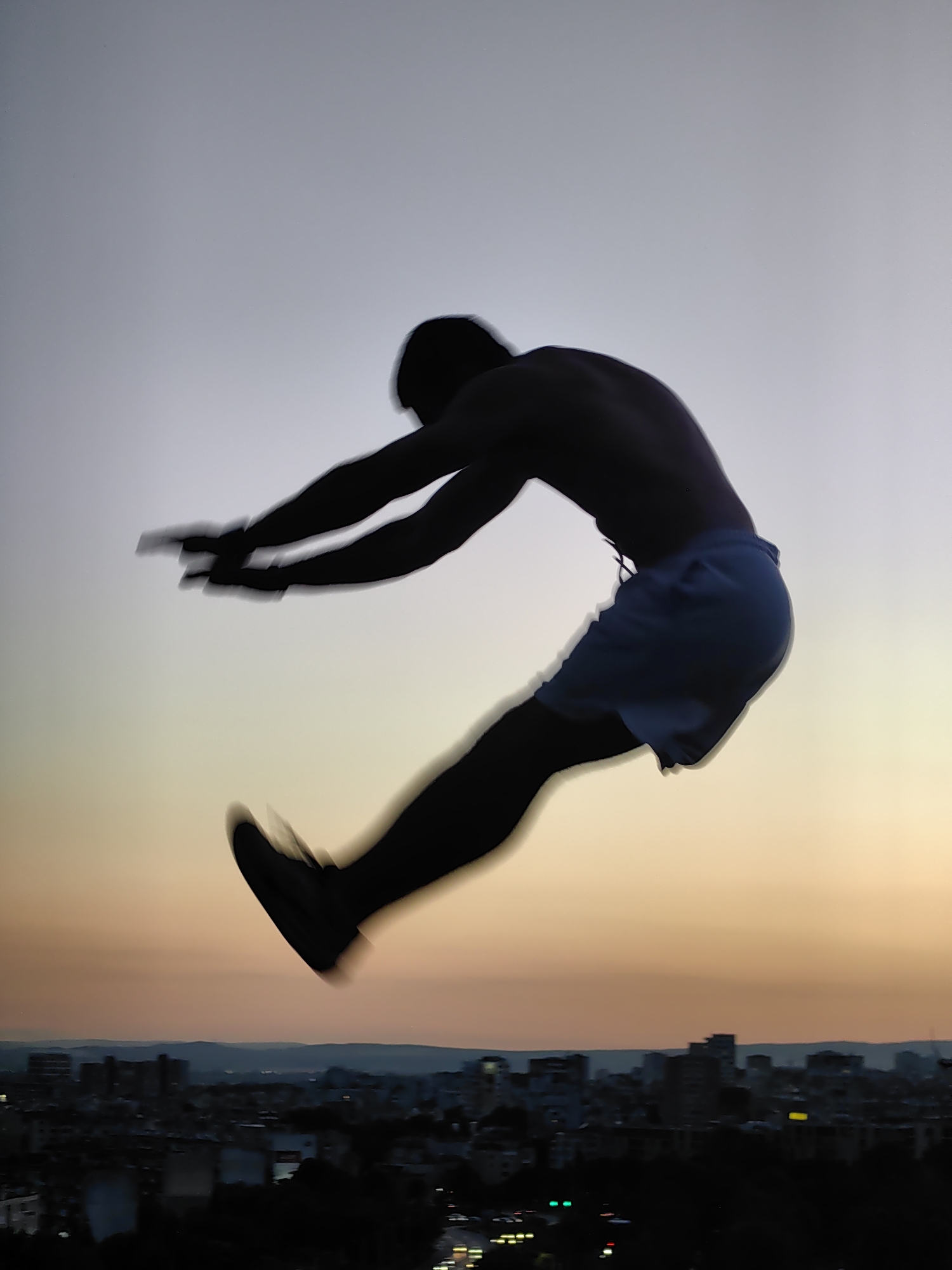 A silhouette of a person in mid-air, with arms extended forward and legs bent backward, is captured against a twilight sky transitioning from pale blue to soft orange. The figure is positioned over a dimly lit cityscape that appears to be Varna, Bulgaria, with buildings and distant hills visible in the background. The motion blur indicates the person is in the process of performing a jump or athletic maneuver.