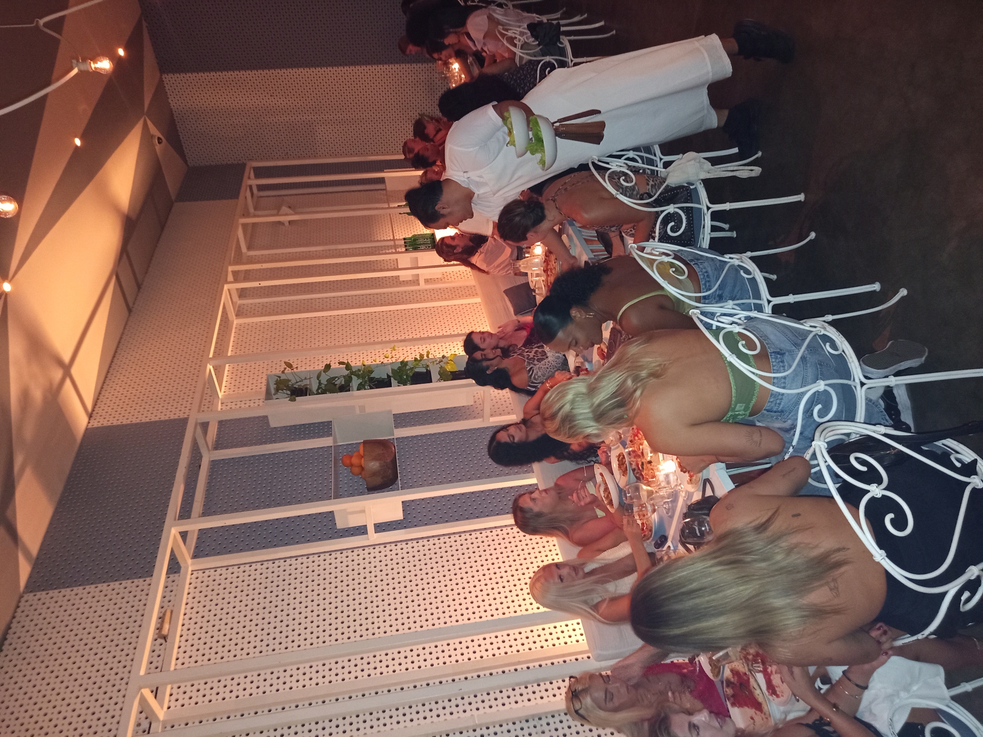 The image captures a group of approximately 10 people seated at a long white table inside what appears to be a restaurant in North Kuta, Indonesia. The table is set with plates of food and glasses, suggesting a meal is in progress. A server dressed in white is seen delivering food to the guests. The interior design includes white metal shelving units with plants and decorative items, as well as a distinctive white textured wall with a pattern of small circles. Overhead, there are strings of lights that cast a warm glow over the scene. The atmosphere is relaxed and convivial, with the guests appearing engaged in conversation and enjoying their meal.