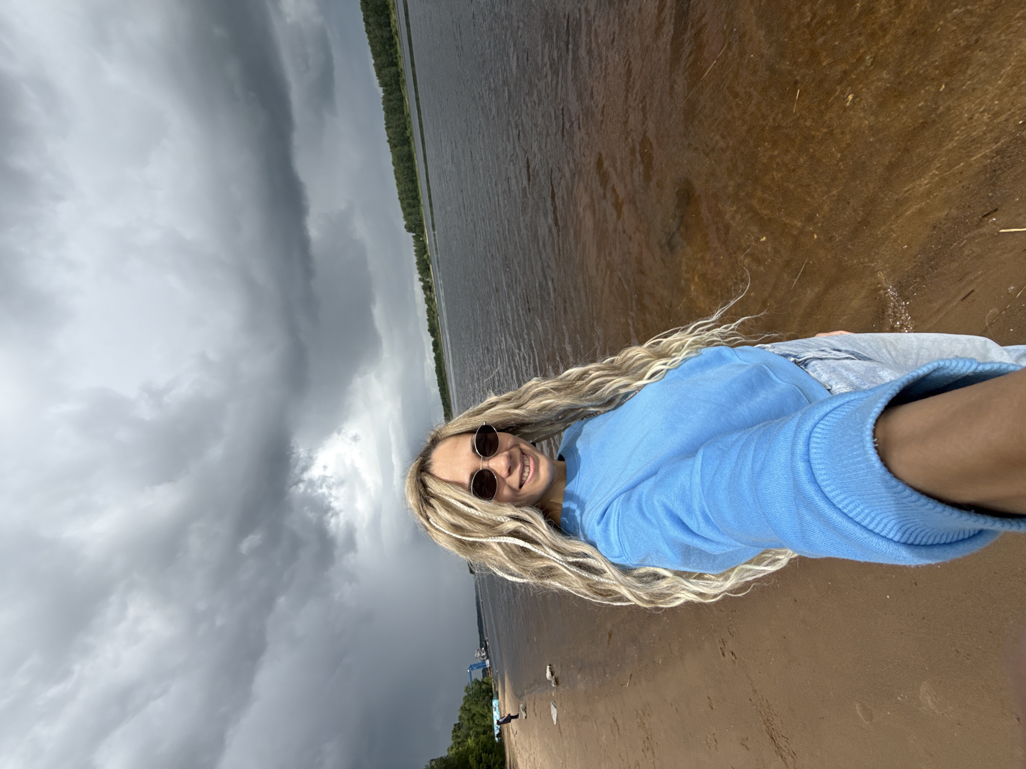 A woman is taking a selfie on a beach. She has long, blonde hair and is wearing sunglasses and a blue sweatshirt. The sky is overcast, and the water is dark. The background shows a treeline across the body of water. The location is likely near the city of Rusanovo, Russia, given the prompt's specifications.