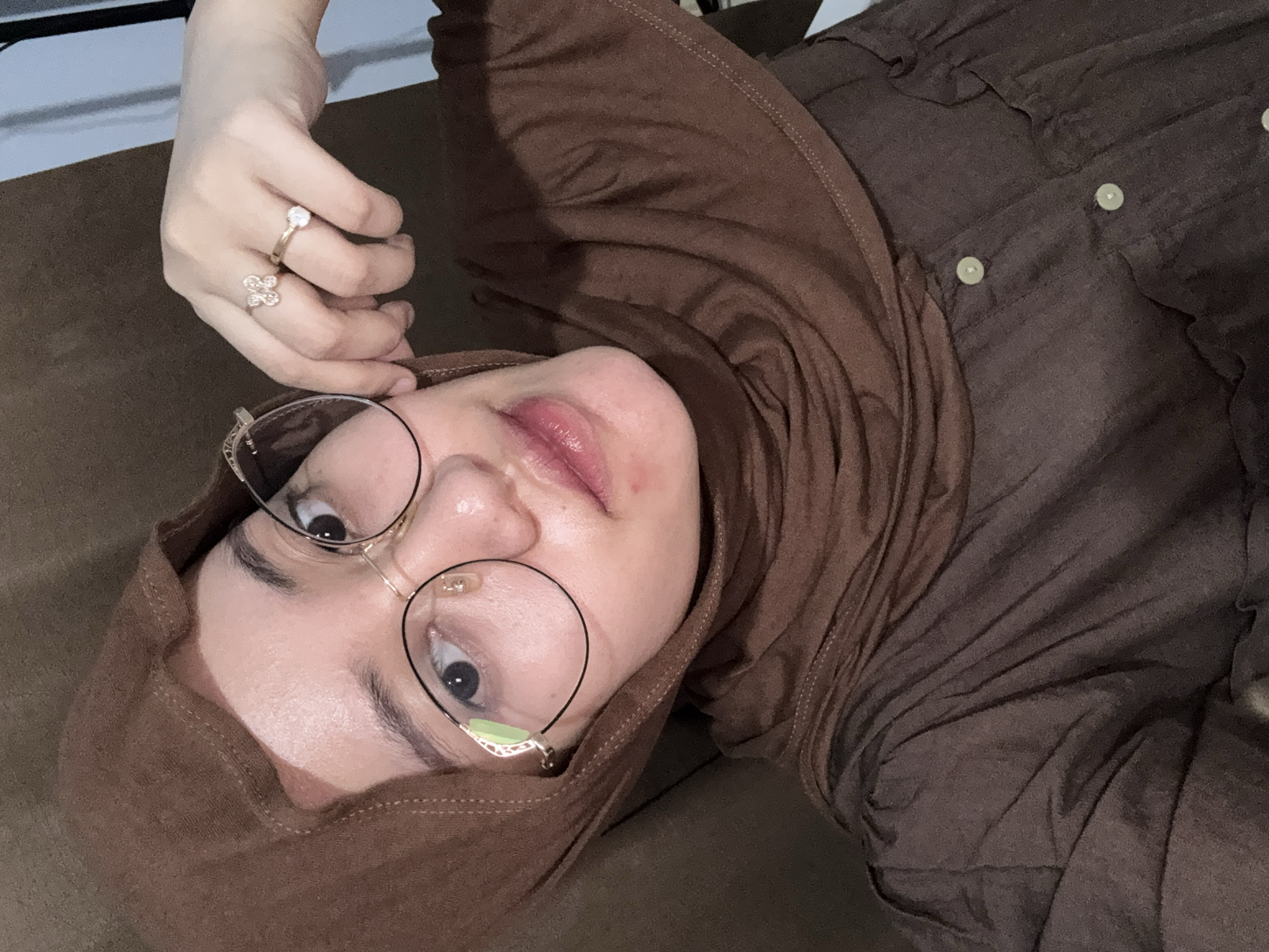 This media file captures a close-up, overhead shot of a person wearing a brown hijab and glasses. The person is lying down, looking upwards at the camera. Their eyes are wide open, and they are wearing a light pink lipstick. On their left hand, they are wearing two rings: one on the ring finger with a large stone and another on the middle finger with a clover-like design. The subject is wearing a brown shirt underneath the hijab. The background appears to be indoors, with a brown surface. The lighting suggests it could be daytime. The setting is Gadingrejo, Indonesia.