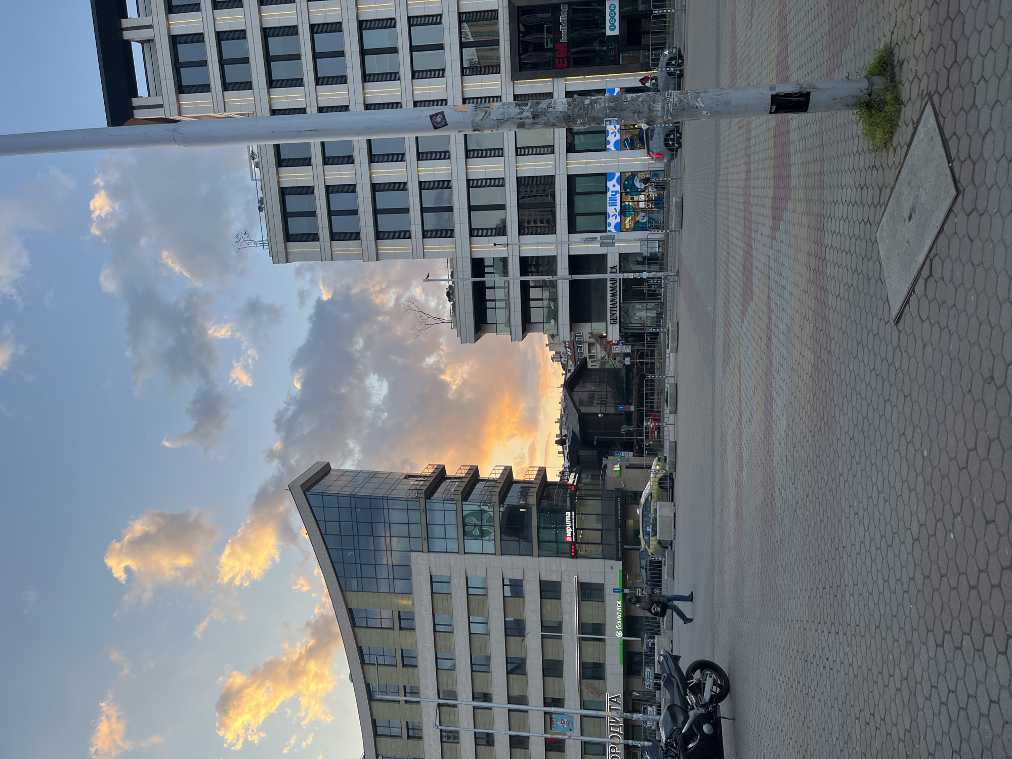 The image shows a cityscape in Sofia, Bulgaria at dusk. In the foreground, a cobblestone pedestrian area stretches into the distance. A grey metal utility pole stands slightly off-center, with exposed wiring and remnants of affixed stickers. Near the pole, grass grows through the paving. The buildings on the left appear modern, constructed of glass and light-colored materials. Signage of businesses are visible. People are walking, while motorcycles are parked near the buildings. The sky is illuminated with the hues of sunset, featuring light-colored clouds.