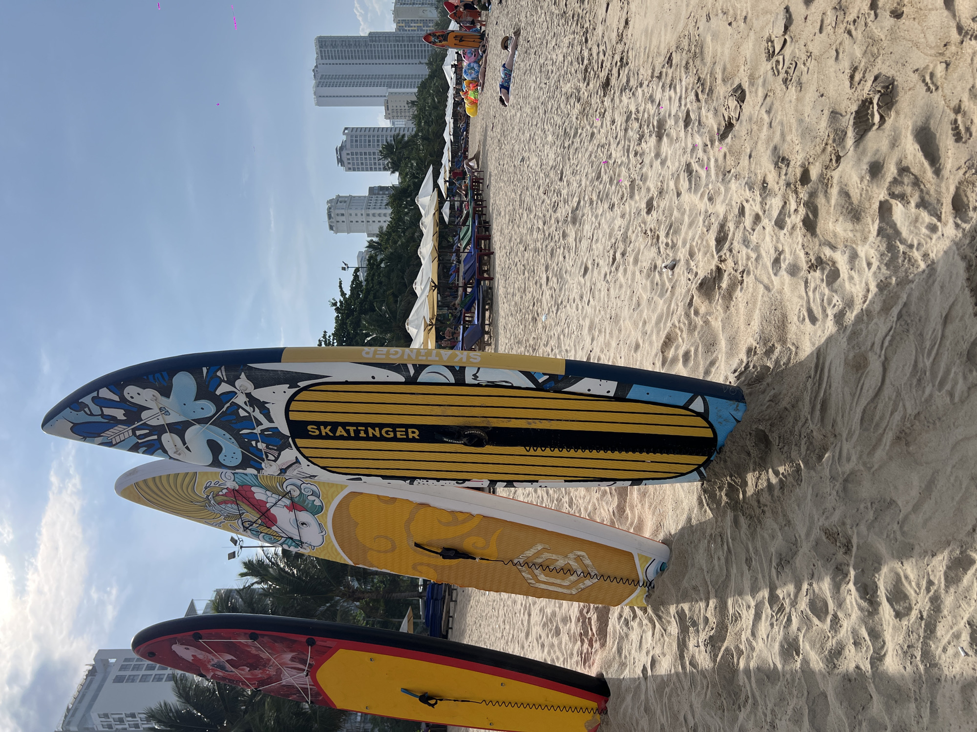 The image displays a sunny beach scene in Nha Trang Ward, Vietnam, during daylight hours. In the immediate foreground, three distinct stand-up paddleboards (SUPs) are propped upright in light brown sand. The leftmost SUP is primarily red and yellow, featuring a graphic of a red koi fish. The middle SUP is yellow and white, adorned with a graphic of a white koi fish, and displays the brand name "SKATINGER." The rightmost SUP is blue, yellow, and black, detailed with abstract designs and prominently features "SKATINGER" printed vertically on its yellow section. Each SUP is equipped with a coiled black leash. Shadows from the SUPs are cast on the textured sand, which exhibits various footprints and ripples.
Extending into the midground is a wide sandy beach occupied by multiple rows of beach infrastructure. These include white sun umbrellas and an assortment of sun loungers in blue, green, and red. Several individuals are present, with some reclining on loungers under umbrellas and others visible further along the beach. One person is distinctly observed lying prone on the sand in the lower right midground. Behind the beach setup, scattered palm trees are visible. The background features a dense urban skyline composed of numerous tall, modern high-rise buildings, consistent with a coastal city. The sky above is largely clear and blue, with minimal scattered white cloud cover. Faint magenta-colored specks are discernible in the upper right quadrant of the sky.