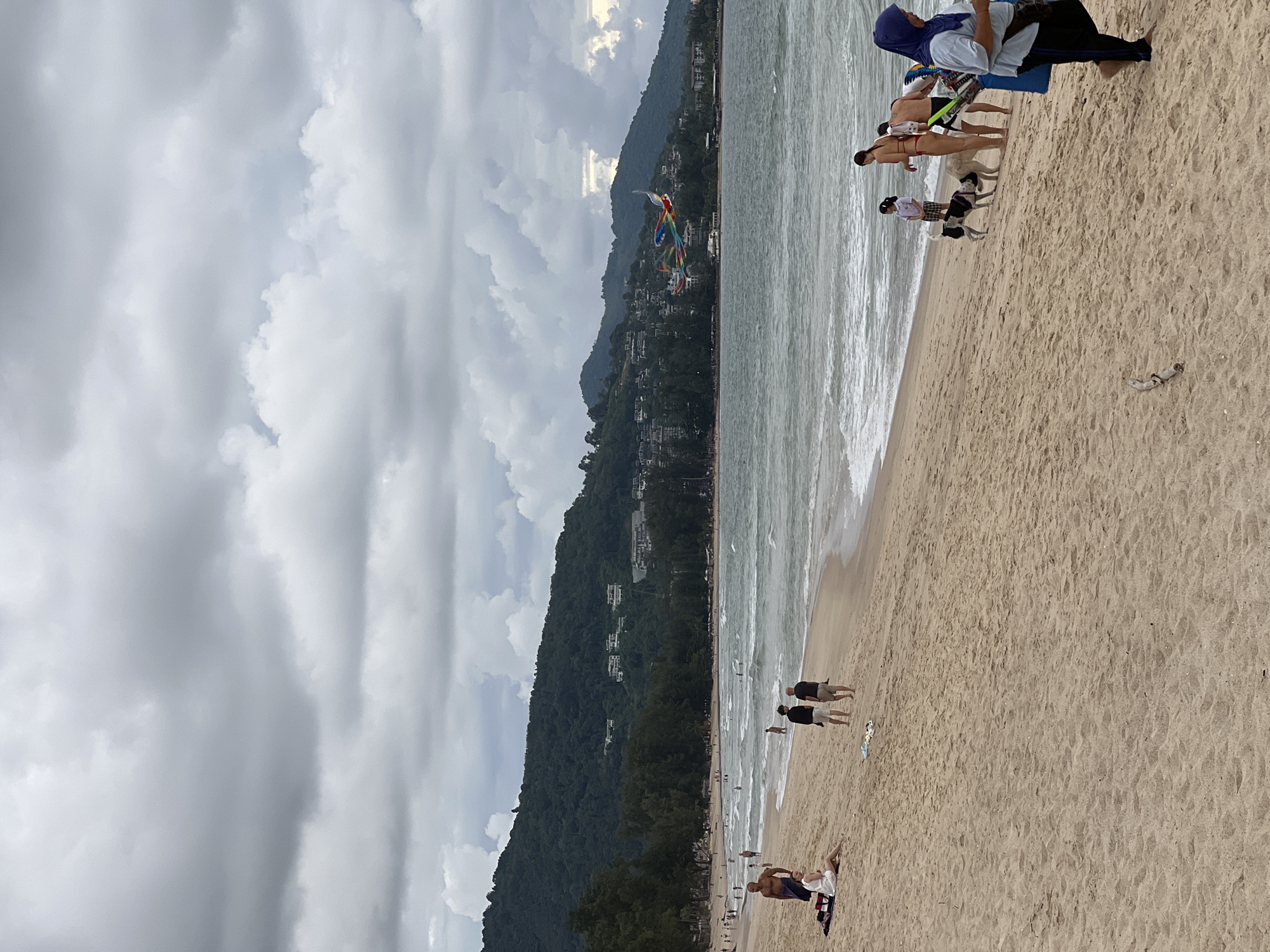 The image captures a beach scene under a cloudy sky. In the foreground, the sandy beach is populated by several individuals and two dogs. A woman in a white dress is sitting on the sand, while two men stand nearby. Further down the beach, a group of people are interacting, including a woman in a bikini, a man, and two children. Two dogs are also present, with one black and white dog near the water and a larger light-colored dog beside it. A woman wearing a blue hijab walks towards the right edge of the frame. In the middle ground, the ocean waves are breaking on the shore, and several people are visible in the water. In the background, a lush green hillside is dotted with buildings, and a colorful kite, possibly in the shape of a bird or dragon, is flying in the sky. The overall atmosphere suggests a somewhat overcast day at a coastal resort area.