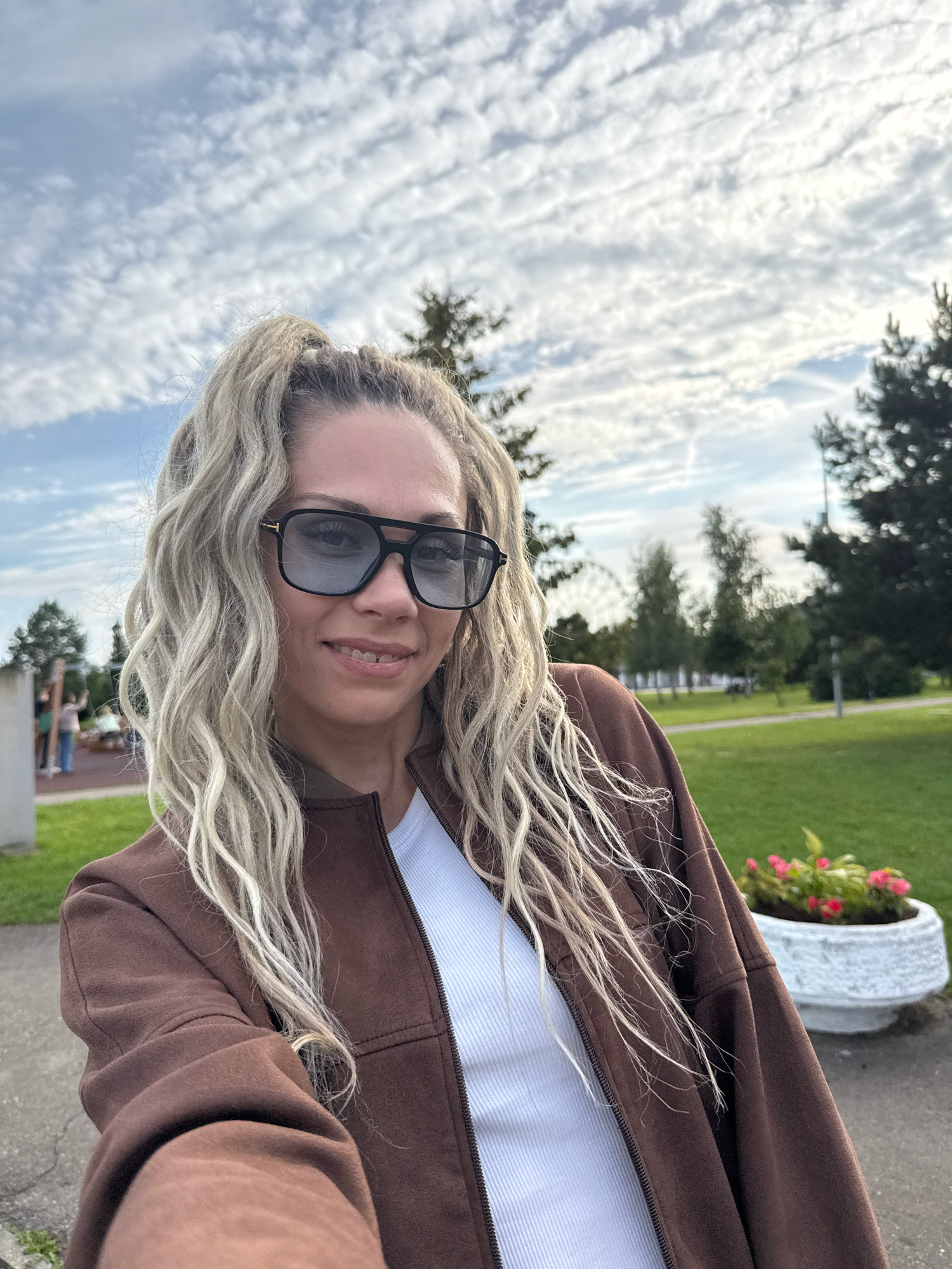 A woman with blonde dreadlocks styled in a high ponytail, wearing large black sunglasses, a white ribbed tank top, and a brown bomber jacket, takes a selfie outdoors. She is smiling and looking at the camera. The background shows a park with green grass, trees, a Ferris wheel in the distance, a playground, and a flower pot with pink flowers. The sky is cloudy. The location is in the city of Zhukovo, Russia.