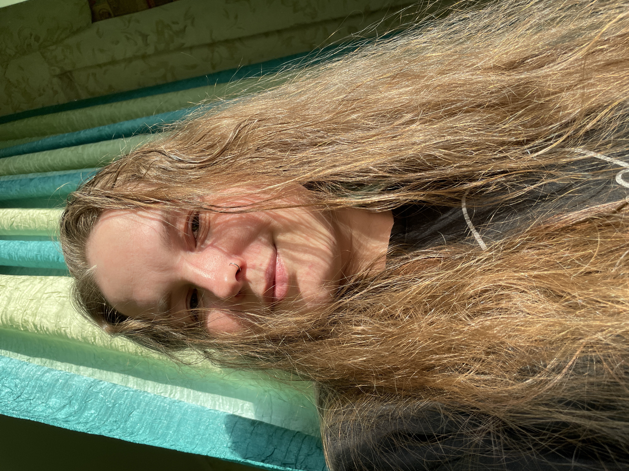 A photograph shows a person with long, light brown or blonde hair lying against a striped surface with alternating bands of light green and teal. The person has fair skin and a small silver ring in their left nostril. The person is wearing a dark-colored shirt or top. The person's hair is spread out around their face. Strong sunlight is present. The location is not discernible from the image. No details indicate this photo was taken in Izhevsk, Russia.
