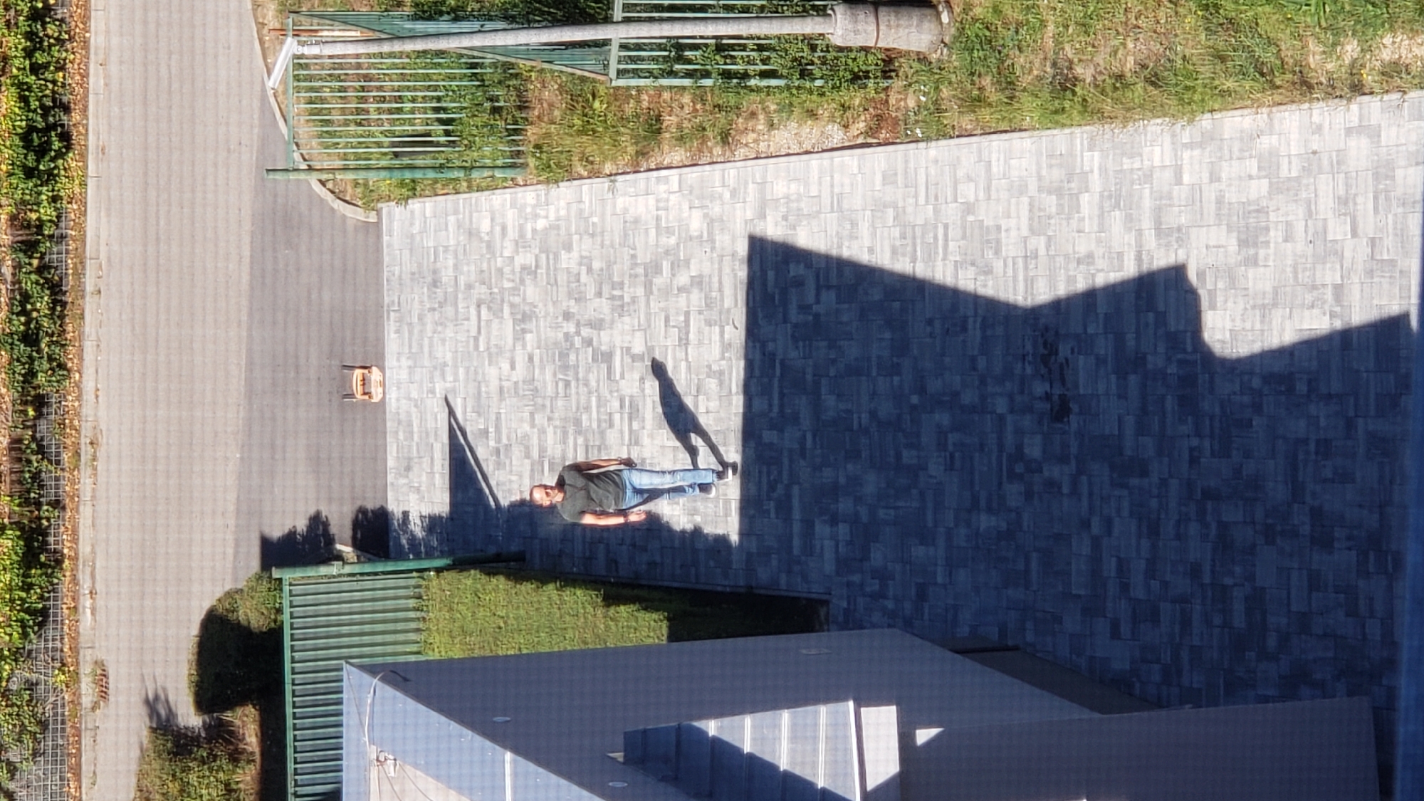 An aerial view shows a man walking on a paved walkway. The walkway is made of gray rectangular pavers arranged in a repeating pattern. To the left of the walkway is a modern building with a light-colored facade and a metal railing. To the right of the walkway is a grassy area bordered by a green metal fence. In the background, beyond the fence, is a street and a row of trees and vegetation. A small, orange, two-wheeled cart is visible on the street in the background. The man is wearing a dark t-shirt, blue jeans, and dark sneakers. He is casting a long shadow to his left, indicating the sun is to his right. The scene appears to be an outdoor public space, possibly a park or a residential area in Prešov, Slovakia, given the context of the location.