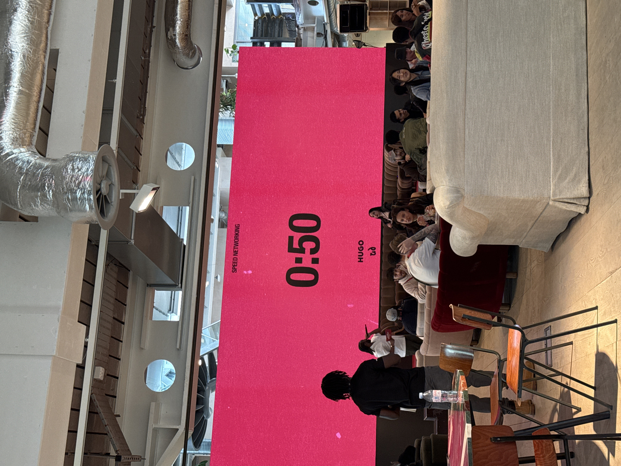 The image shows an indoor scene in London, United Kingdom. A group of people sit on a couch and chairs, presumably participating in a "Speed Networking" event, as indicated by the large pink screen in the background displaying "0:50" and "HUGO." A person is standing near the screen, possibly managing the event. The room features exposed ductwork and a large window. Other chairs and a table are visible in the foreground.