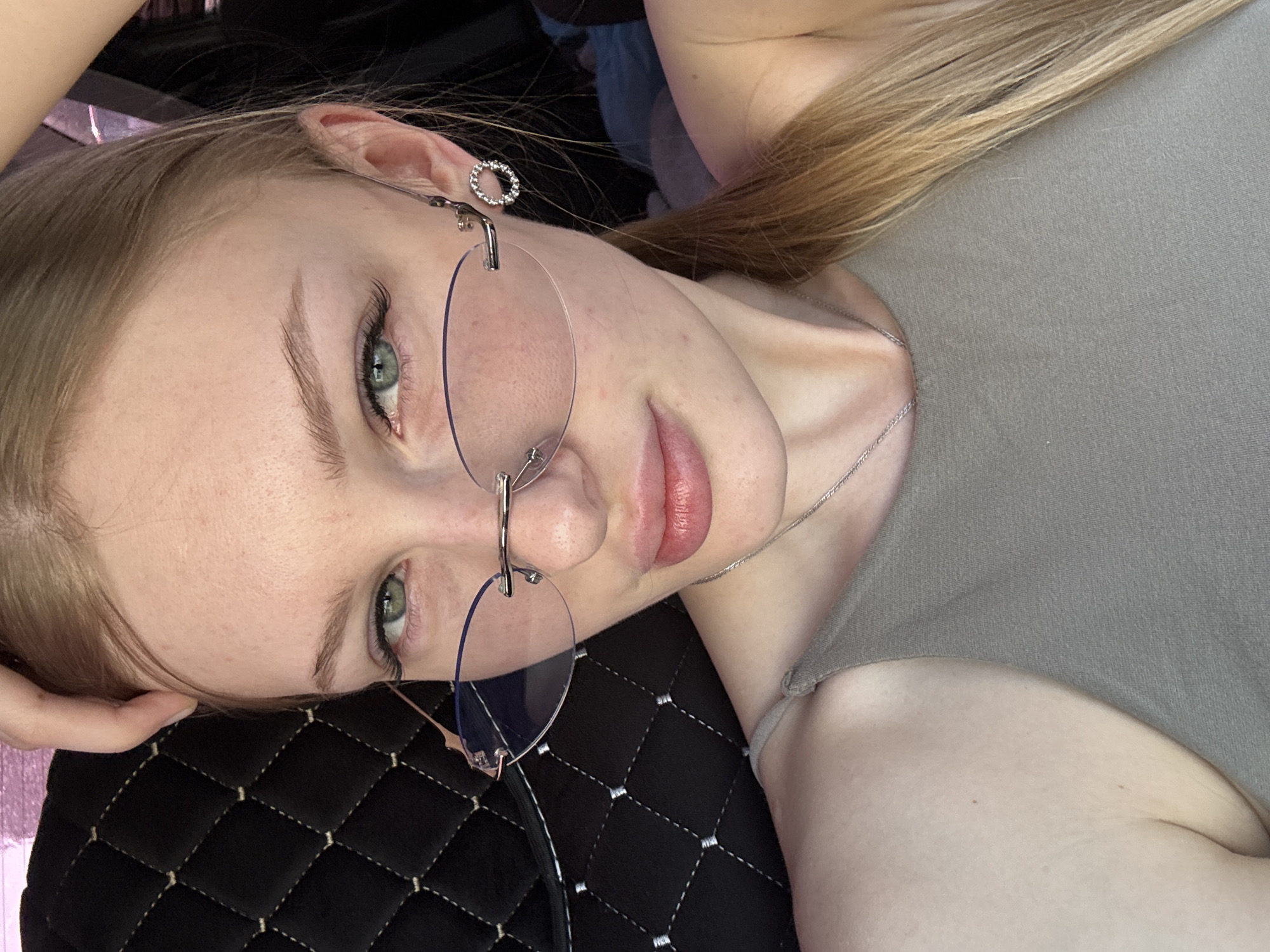 A young woman is pictured in a close-up, slightly tilted shot. She has light skin, blonde hair, and is wearing rimless glasses and a circular earring in her left ear. Her eyes are open and looking towards the viewer. She is wearing a light-colored top and a thin silver necklace. The background consists of a black quilted surface and glimpses of what appears to be car interior elements. The image was taken in Perm, Russia.
