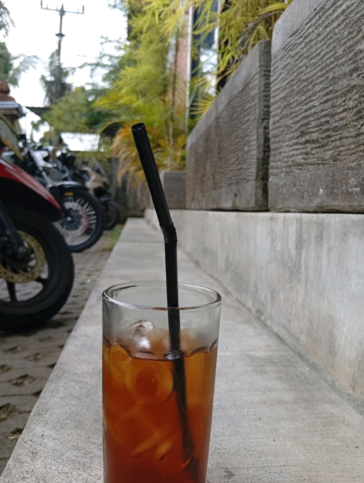 A close-up, eye-level shot focuses on a tall, clear glass filled with iced tea and a black straw. The glass is positioned on a concrete ledge, with a line of motorcycles blurred in the background to the left and textured stone blocks to the right. The iced tea has a rich amber color, with visible ice cubes and condensation on the glass. The straw, a dark, ribbed cylinder, extends from the drink towards the top of the frame. In the background, out of focus, are glimpses of greenery, a utility pole, and the edge of a building. The scene suggests an outdoor setting, possibly a cafe or street-side seating area in a tropical urban environment like Ubud District, Indonesia, given the presence of motorcycles, lush foliage, and architectural elements. No people are visible.