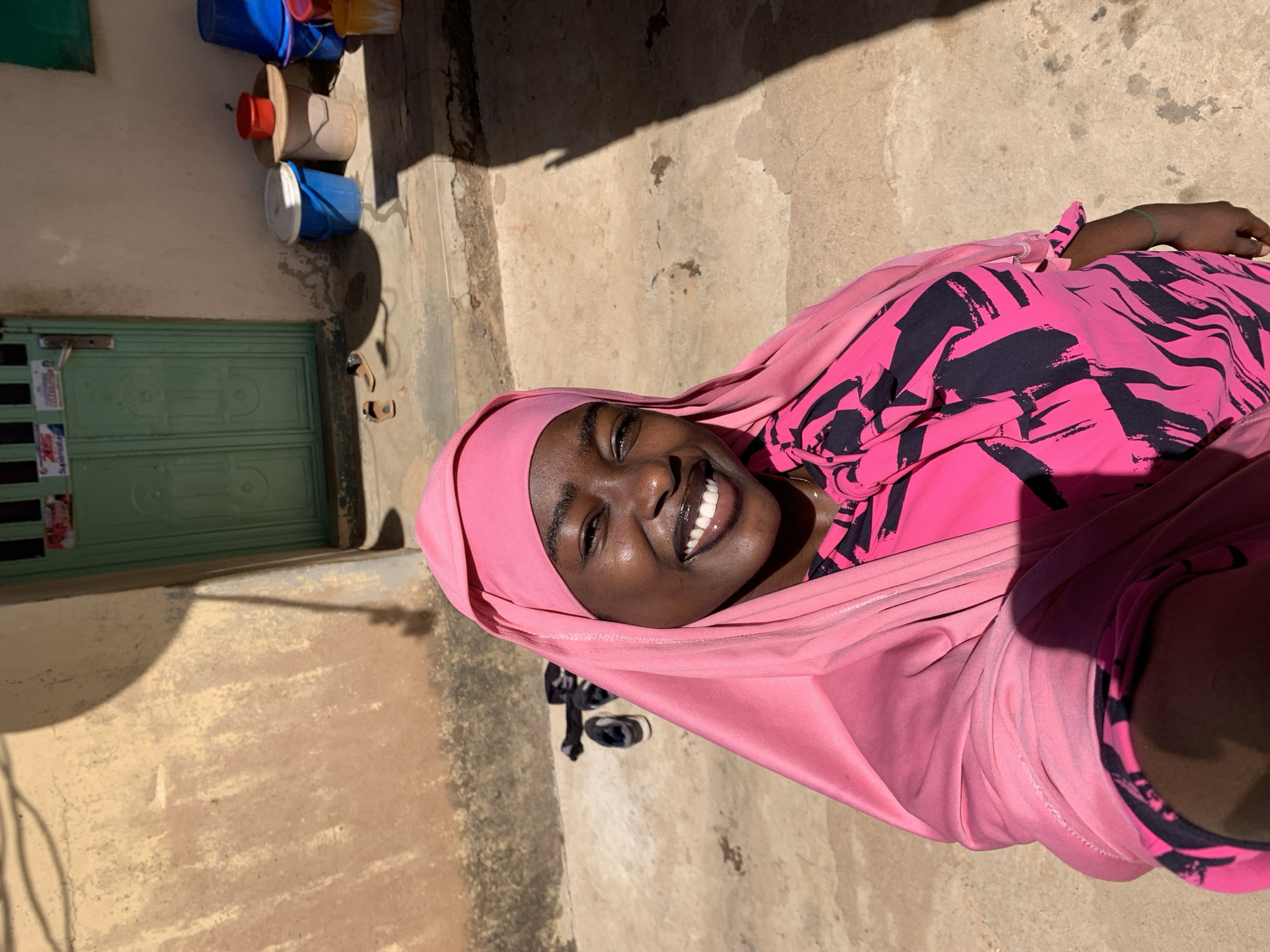 A young woman is lying down on a textured surface of tan and gray concrete, with a tan wall and green double door visible in the background. She is wearing a pink headscarf and a pink top with a black pattern. She smiles at the camera with her eyes open. To the upper left of the image are some buckets stacked on the ground near the green door. The scene is brightly lit with natural light. The setting appears to be outdoors in the city of Rafin Par, Nigeria.