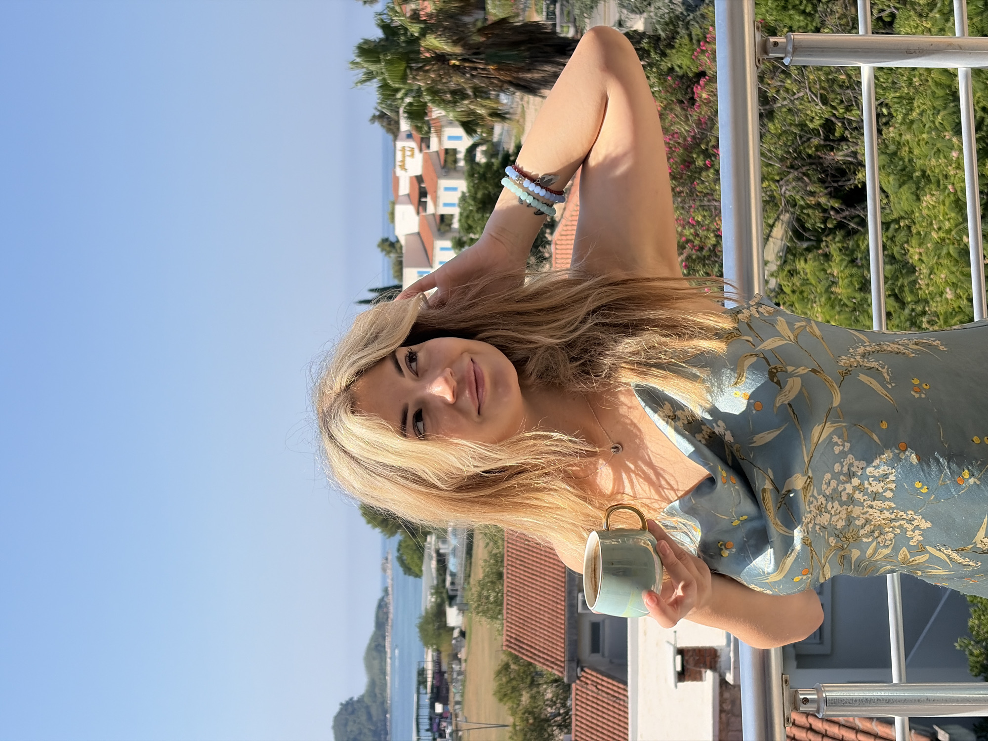 A young woman with blonde hair stands on a balcony overlooking the city of Budva, Montenegro. She is holding a teal mug with a gold handle in her left hand and her right hand is raised to her hair. She wears a light blue slip dress with a floral pattern. The background shows buildings with red-tiled roofs, palm trees, and the Adriatic Sea. The sky is clear and blue.