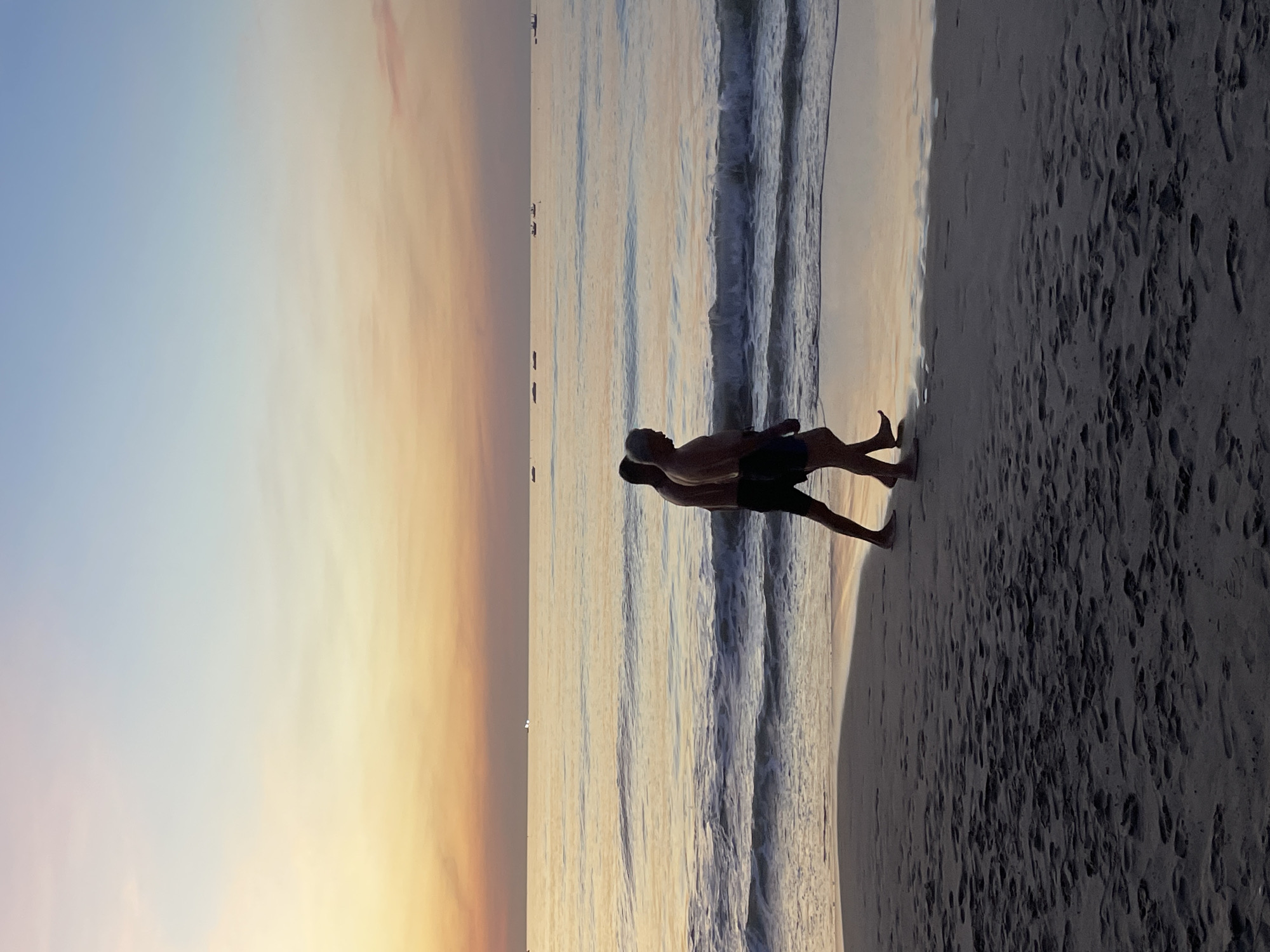 The image depicts a serene beach scene at what appears to be sunset or sunrise, given the soft, warm hues in the sky transitioning from blue to orange. Two people are visible on the sandy shore. One person, silhouetted and wearing dark shorts, is lying down or resting on the sand. Another person, also silhouetted, is standing or walking, leaning over the first person as if to embrace or assist them. Gentle waves are rolling onto the shore, creating a rhythmic pattern on the water's surface. The beach is marked with numerous footprints in the sand, indicating recent activity. In the distance, several small boats are scattered across the water, and a faint line of land or structures can be seen on the horizon. The overall mood is peaceful and intimate, suggesting a shared moment between the two individuals in a tranquil natural setting. The location is identified as Đà Nẵng, Vietnam. No text is visible in the image.