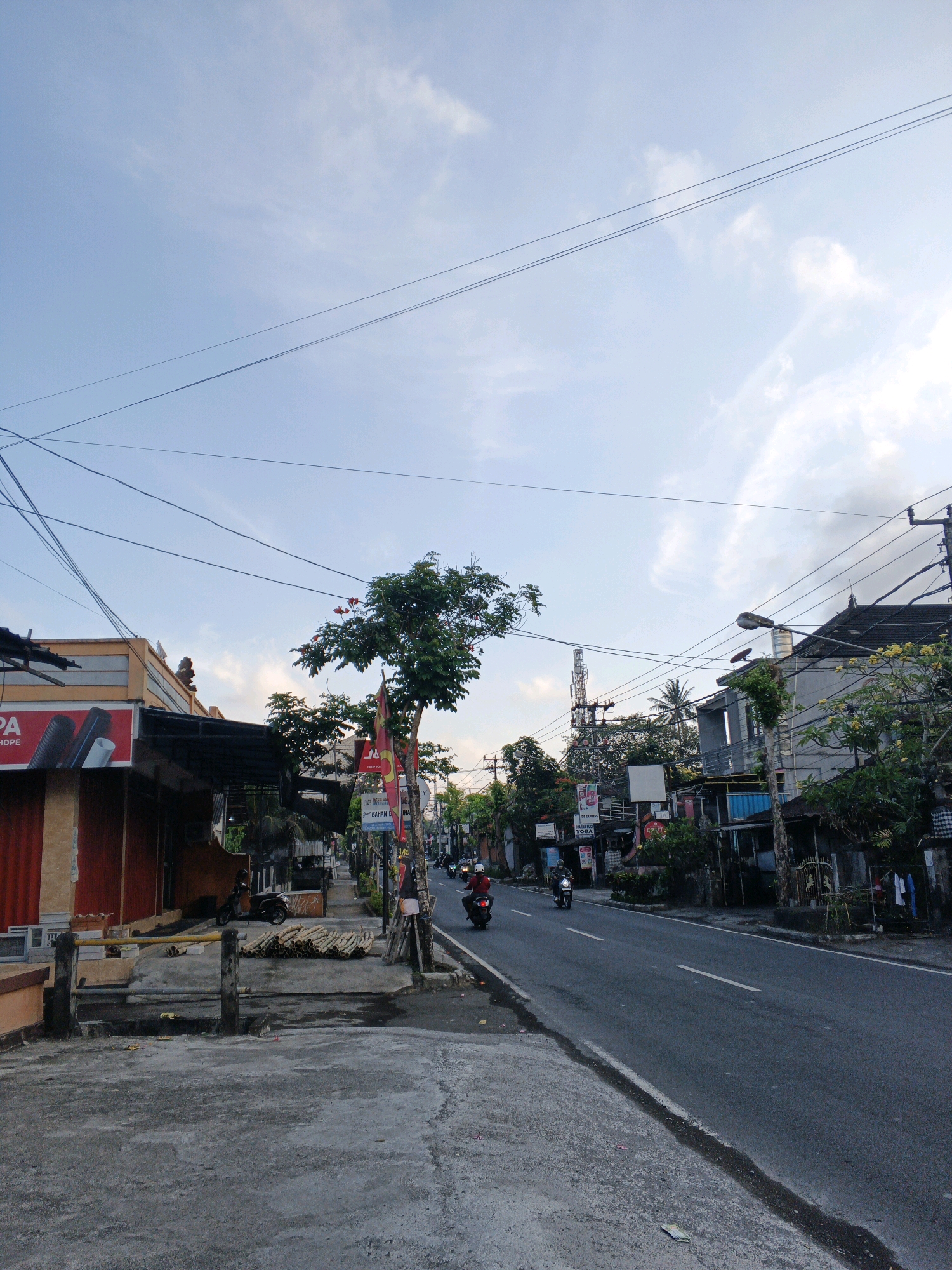 A street scene in Abiansemal, Indonesia, shows a road with light traffic under a partly cloudy sky. The road is asphalt with white lane markings. On the left side of the road, there are buildings with awnings and shop fronts. One building has an orange facade and a sign with the letters "PA" visible. Piles of cylindrical materials, possibly bamboo or logs, are stacked on the sidewalk. Further down the street, there are more buildings, some with visible roofs and balconies, and trees with yellow flowers. A cell tower is visible in the background. Several motorcycles are present on the road, with riders visible on some. The overall environment suggests a commercial or residential street in a tropical or subtropical climate.