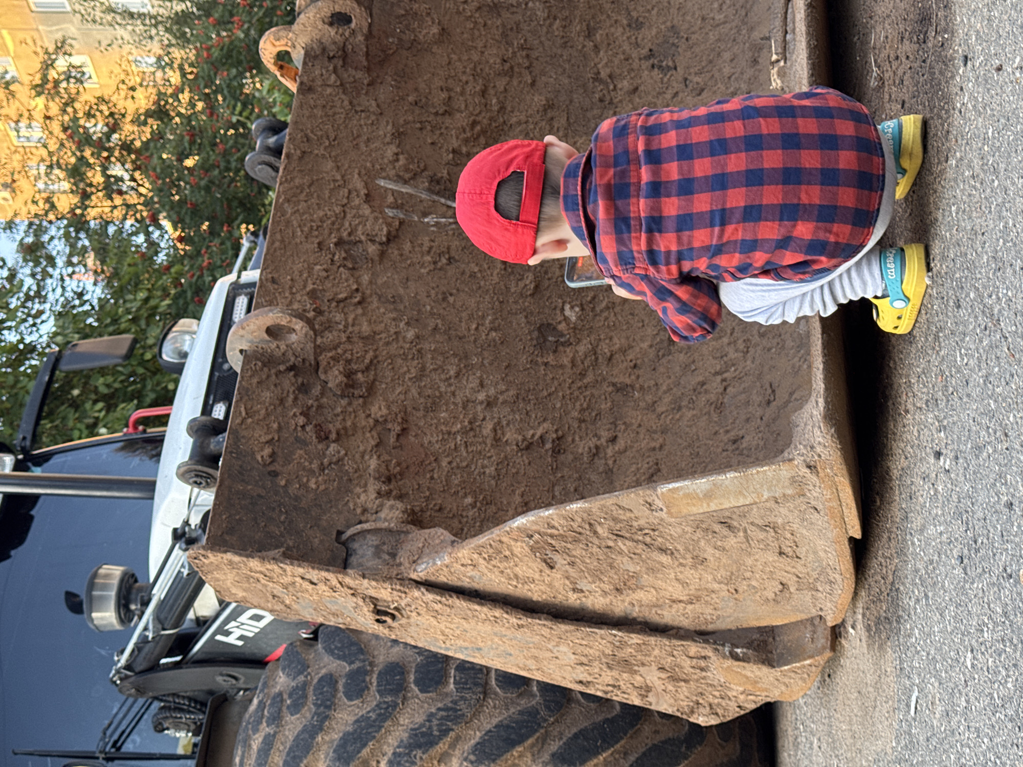 A young child wearing a red baseball cap and a red and blue plaid shirt is crouching on asphalt, facing away from the camera. The child is holding a smartphone and appears to be looking at it. The child's feet are clad in yellow Crocs. Behind the child is the large, dirt-covered bucket of a heavy machinery vehicle, likely a loader. The bucket is rough and appears to have been used for excavation or moving earth. Parts of the vehicle's tire and chassis are visible to the left of the bucket. In the background, there is a building with windows and a tree with red berries, bathed in the warm light of what appears to be late afternoon or early evening. The overall scene suggests an urban environment, possibly a construction site or a street in Izhevsk, Russia, given the context.
