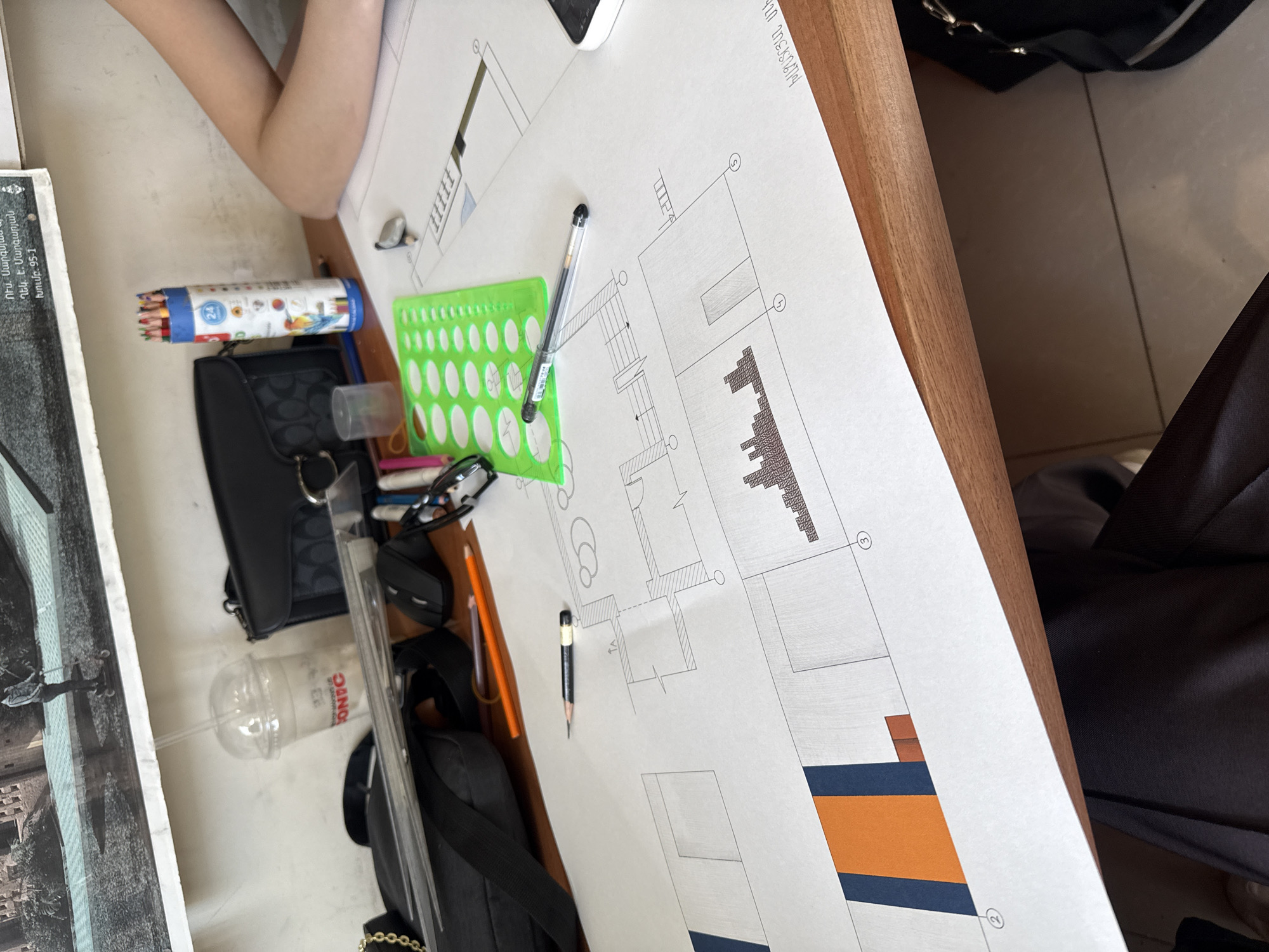 The image captures a desk with various objects related to architectural drawing. A large sheet of paper displays a partially completed architectural plan, featuring lines, dimensions, and symbols indicating walls, doors, and other structural elements. A pen and a pencil rest on the plan, alongside a green stencil. A black bag, a container of colored pencils, a measuring tool, eyeglasses, and a plastic cup with a straw are also visible on the desk, suggesting the ongoing work of a designer or architect.
The text on the drawing, which includes "ԽԱՉԱՏՐՅԱՆ ՍՆԷ" and "Ղեկ. Է. Մարգարյան," indicates this is a project associated with individuals named Khachatryan Sne and E. Margaryan. The text "ՈՒս. Սարգ," and "ใบทเปρ. 95-1" suggests it may be related to a university project, potentially at Yerevan State University of Architecture and Construction (YSUAC), given the context of architectural drawings and the possible location in Yerevan, Armenia.