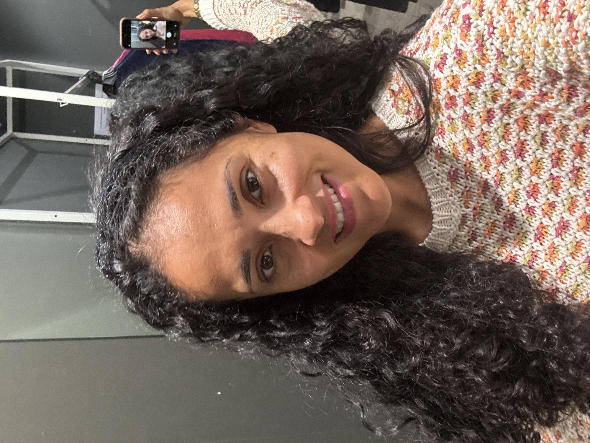 An indoor, eye-level shot captures a woman taking a selfie, rotated counter-clockwise. The main subject is a woman with dark, abundant, curly hair that frames her face and extends beyond her shoulders. She is looking directly at the camera with a broad, genuine smile, revealing her teeth. She wears a light-colored, knitted top featuring a vibrant, intricate pattern of small, interlocking shapes in shades of pink, orange, yellow, and green.
In the upper left corner of the image, her right hand is visible, holding a dark-cased smartphone. The phone's screen displays a clear reflection of her face, indicating she is actively taking a selfie. The background suggests an indoor environment; the lower half of the frame shows a dark, featureless surface, possibly a floor or wall. In the upper background, behind her head and the phone, there's a glimpse of a light-colored, possibly metallic, structural element and some darker, indistinct shapes. The lighting appears soft and even, highlighting her facial features. No specific time of day or weather conditions can be determined from the image. The photo provides no specific textual information but is set in Luxembourg, Luxembourg.