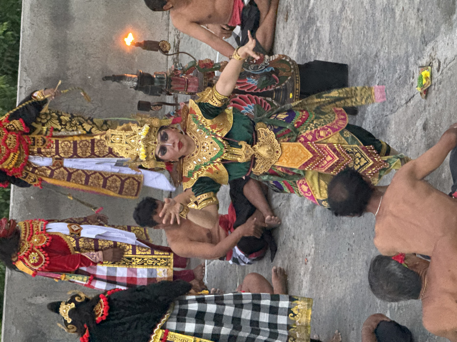 This image captures a traditional Balinese performance in an outdoor setting within Uluwatu, Indonesia. The ground is a light grey, textured concrete or paved surface.
A central female performer is depicted mid-dance, facing forward with both arms extended outwards, hands posed. She wears an elaborate, multi-layered costume featuring a dark green top with extensive gold ornamentation, a prominent golden headdress, and a lower garment with intricate gold, red, green, and pink patterns. Her face is made up with pronounced eyeliner and red lipstick.
To her left and slightly behind, a shirtless male is seated on the ground, wearing dark shorts and a patterned sarong, with his right hand obscuring part of his face. Behind him, a costumed male figure stands, wearing a red, fanged mask, a red and gold jacket, and a white and black checkered sarong.
To the right of the central dancer, a tall, dark, multi-tiered object, likely a ceremonial lamp, is present with a visible flame at its apex. Further to the right, another shirtless male is seated on the ground, partially visible. In the background, another elaborately costumed figure stands, wearing dark and gold patterned attire over a white garment.
In the immediate foreground, the backs of two shirtless males are visible, seated on the ground and facing towards the performance. One individual has a red flower tucked behind his left ear. On the ground near the central dancer's foot, a small Balinese offering, identified as a *canang sari*, is visible. The background consists of a plain, grey concrete wall or structure.
