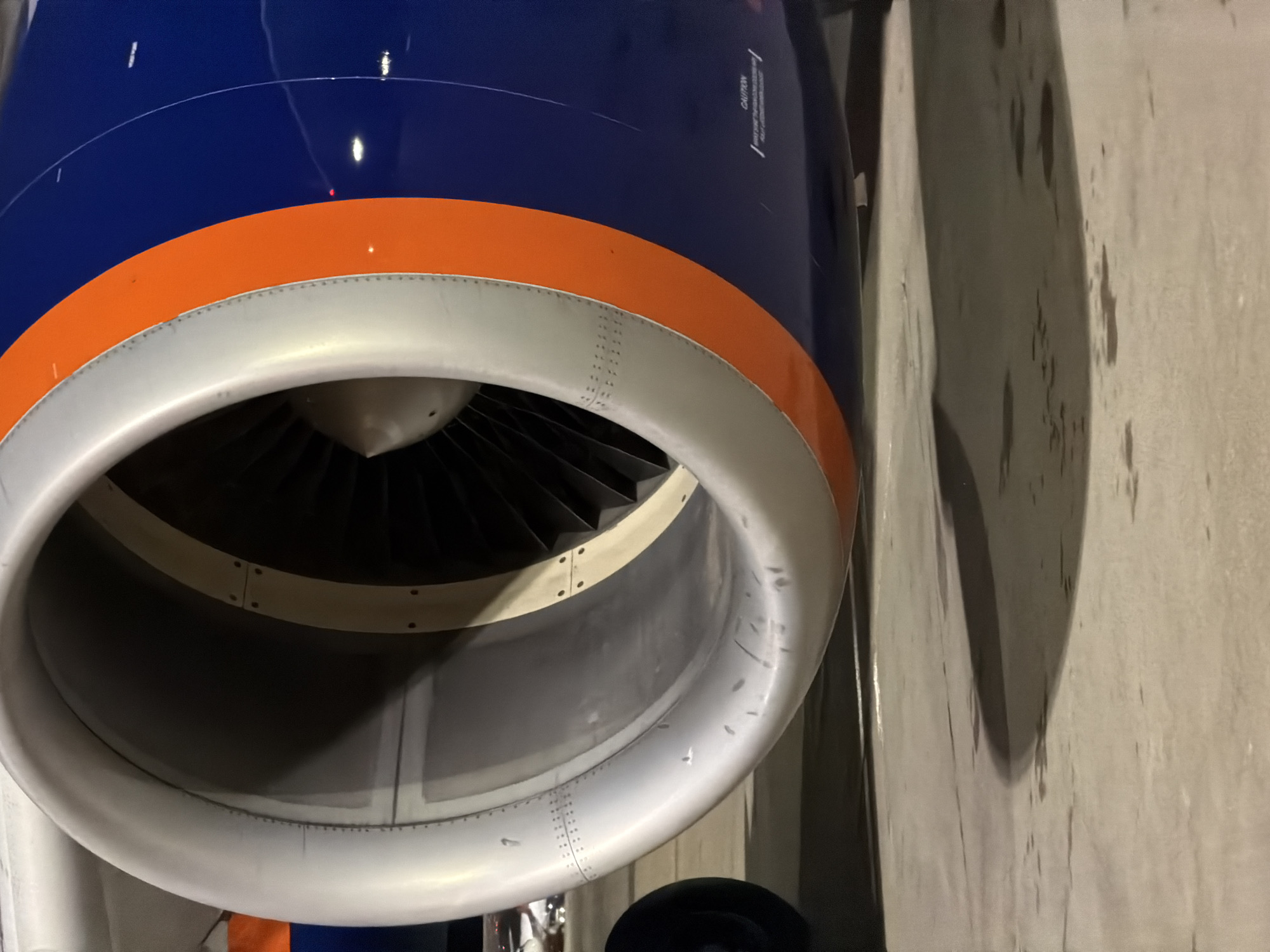 The image shows a close-up, eye-level view of a jet engine from an aircraft, likely at an airport. The engine cowling is predominantly silver with a bright orange stripe that circles its circumference, adjacent to a section of deep blue fuselage. Inside the engine intake, fan blades are visible, shrouded in darkness. The ground beneath the engine appears to be asphalt or concrete, wet in places, with a distinct shadow cast by the engine. In the distance, out of focus, lights and parts of another aircraft or ground equipment can be seen. The scene suggests the aircraft is stationary, possibly parked at a gate or on a tarmac, with no people or active operations immediately apparent. This image does not provide any information to determine its location in the city of Saint Petersburg, Russia.