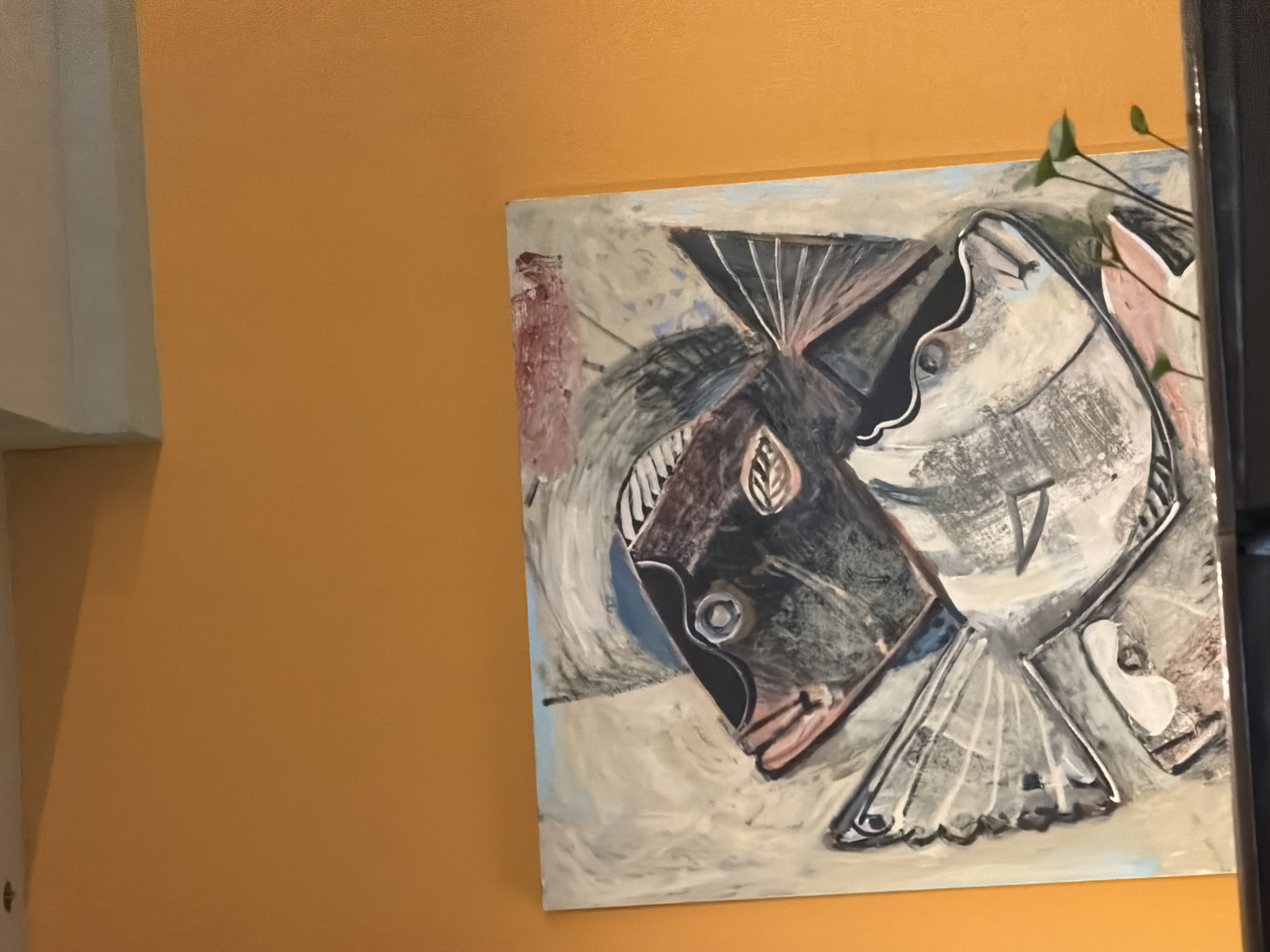 The image shows an abstract painting of two fish hanging on a mustard-colored wall. The painting features two fish depicted in shades of gray, black, and white with accents of red and blue. The painting appears to be oil or acrylic on canvas. The style is abstract and somewhat cubist. A plant with green leaves is partially visible to the right of the painting. A white vertical structure is visible on the left of the frame. The image was taken indoors. The location is unknown and cannot be determined from the image. There is no information available about the city of Plovdiv, Bulgaria.
