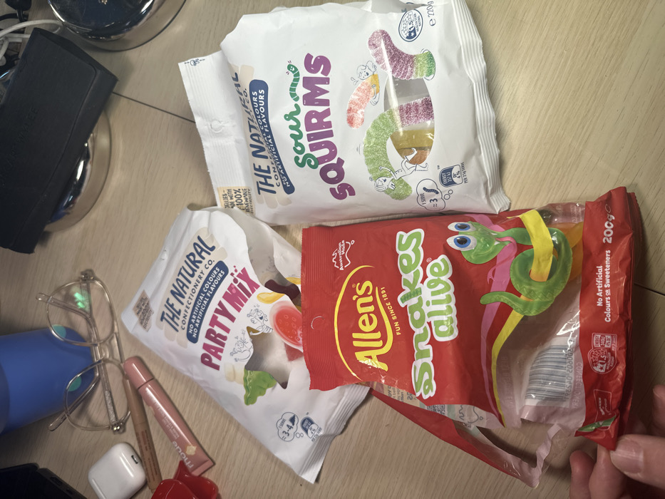 On a light-colored wooden desktop surface in London, United Kingdom, three packages of confectionery and several personal items are arranged. A red, partially open bag of "Allen's Snakes alive" (200g) is held at its bottom edge by a hand, revealing multi-colored snake-shaped gummies inside. Adjacent to it, a white, sealed bag of "The Natural Confectionery Co. Sour Squirmis" (220g) displays sour worm-shaped gummies. Behind these, a third white bag of "The Natural Confectionery Co. Party Mix" is visible. Other items on the desk include a pair of clear-framed eyeglasses, a white Apple AirPods case, a pink "mode" brand lip gloss, a brown cosmetic pencil, a partially visible blue water bottle, and a black object resting on a silver circular base. Portions of other metallic objects and cables are discernible in the background.