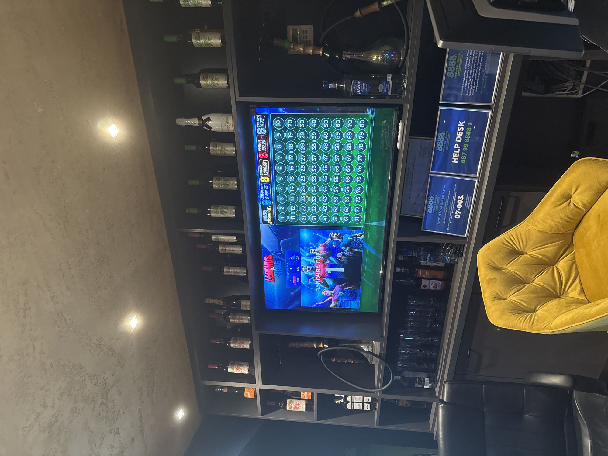 The image shows an indoor space, possibly a bar or lounge, featuring a large screen displaying a soccer-themed casino game with numbered slots. The screen is surrounded by shelves filled with wine bottles and various liquors. To the right of the screen are signs with the number "8888" and the phrase "HELP DESK" along with a phone number. A yellow chair is partially visible in the lower right, suggesting seating for patrons. Below the chair is a black leather chair. A hookah sits on one of the shelves. The lighting in the room is dim, with spotlights on the wall. The image has the characteristics of being taken in Burgas, Bulgaria, given that the OCR detected Bulgarian language on the screen and signs.