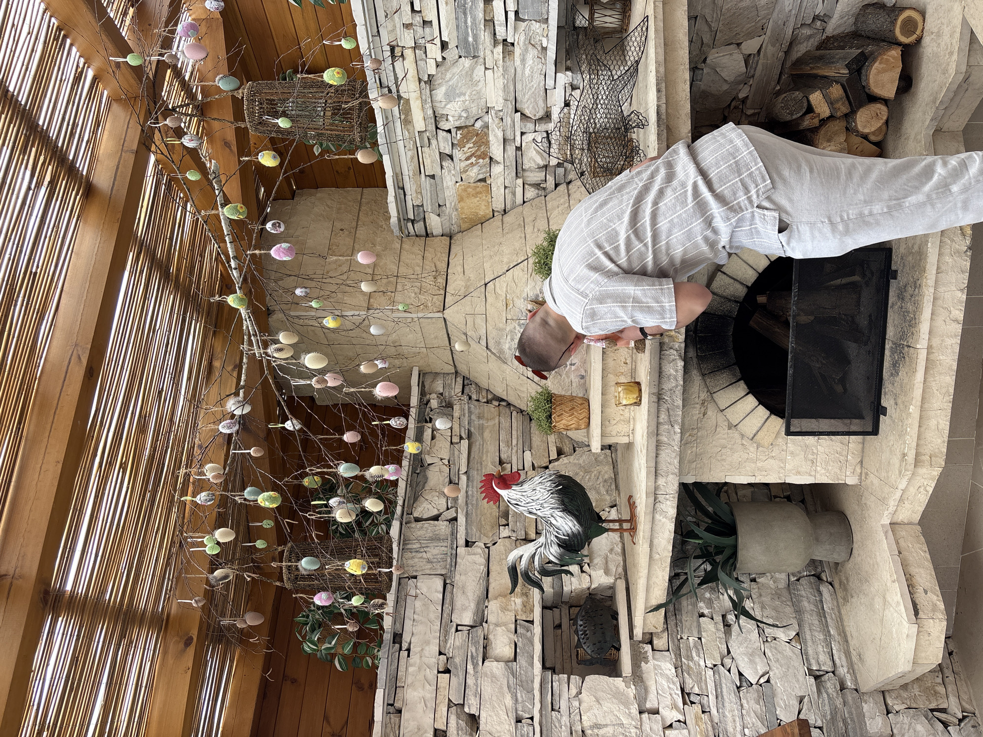 The image shows an interior space, likely in a residence, potentially in Koprivlen, Bulgaria given the request. A man dressed in light-colored linen clothing is leaning over a fireplace, examining the contents within. The fireplace is constructed of light stone and has a black metal grate. A stack of firewood is visible to the right of the fireplace opening. A large stone wall features a rooster sculpture and a hanging basket with greenery. To the left, a decorative tree with Easter eggs hangs, set against a background of wooden slats. The overall setting suggests a rustic and potentially artistic or decorative environment.
