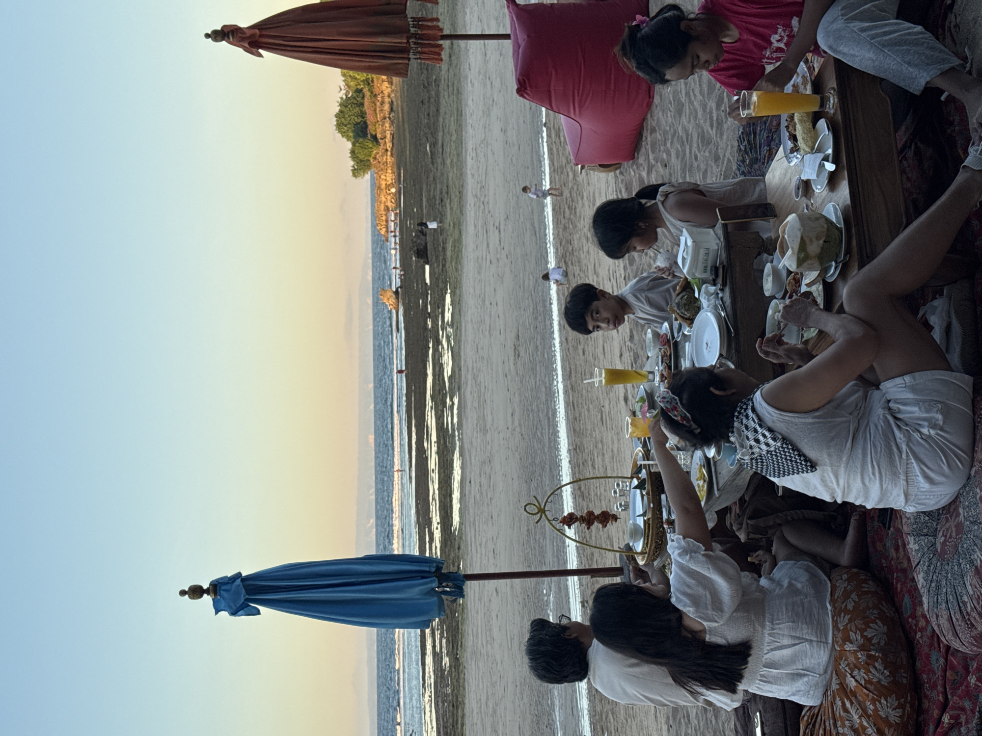 The image shows a group of people gathered around a table on a beach, likely enjoying a meal or gathering. The location appears to be Kuta Selatan, Indonesia, given the coastal landscape and beach setting. There are two umbrellas, one blue and one orange. In the background, the ocean and the horizon are visible. The group around the table includes individuals of varying ages. They are seated on cushions. The table is laden with food and drinks. Some individuals are eating or drinking, while others seem to be engaged in conversation. The overall atmosphere is relaxed.