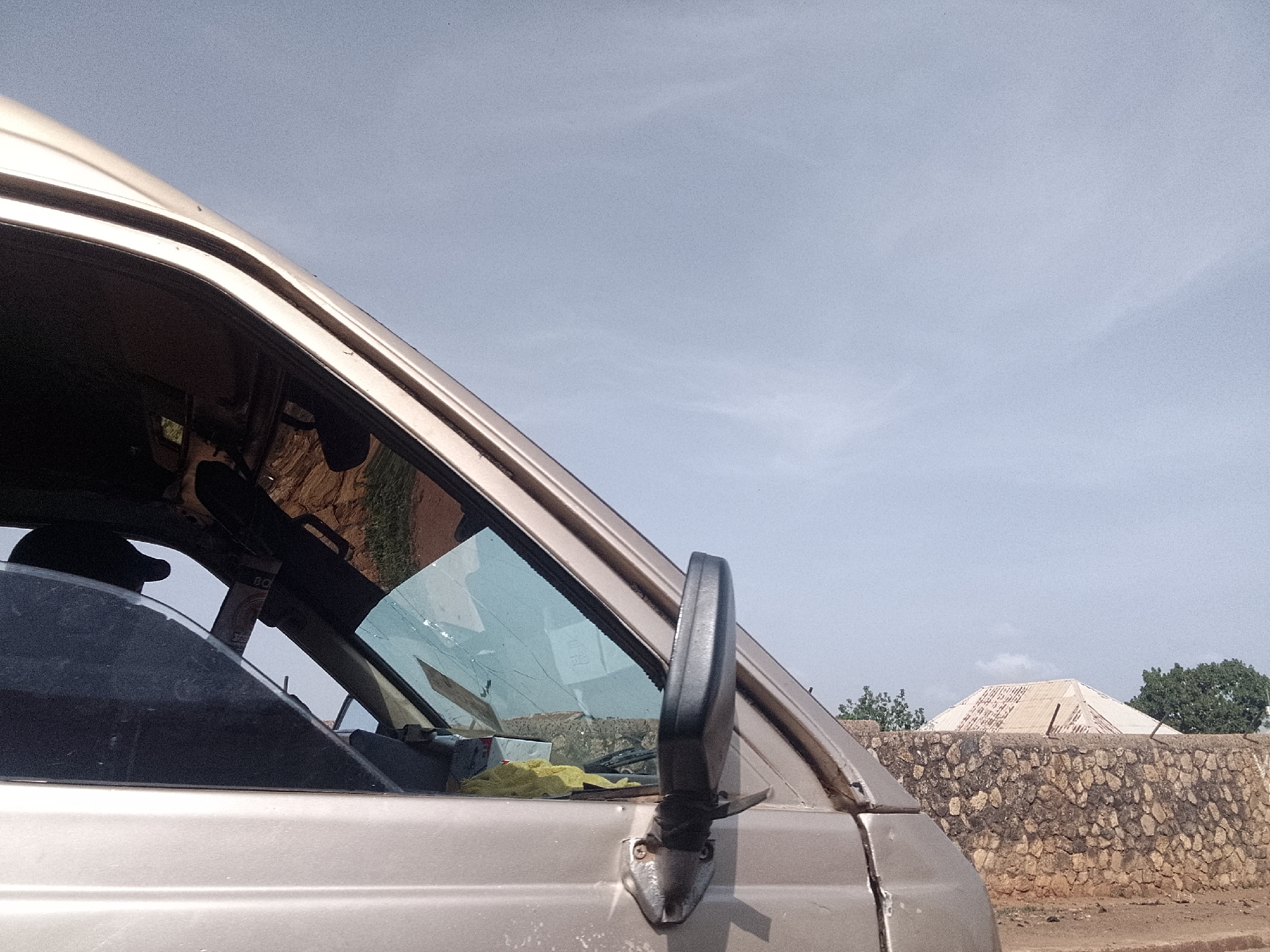 The image presents a view from inside or very close to a vehicle, likely a van or a truck, parked on a street in Bukuru, Nigeria. The vehicle’s side mirror is visible, along with a partial view of the dashboard and front window. Outside, a stone wall topped with barbed wire is visible, and behind the wall, there’s a building with a distinctive pyramid-shaped roof. The sky is overcast, and there are trees in the background.