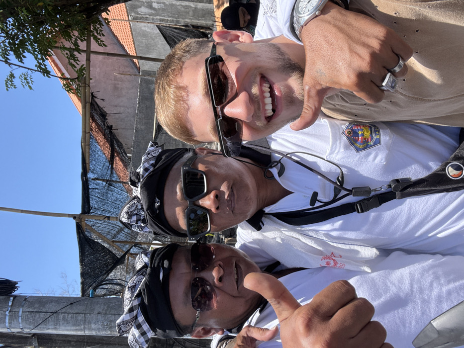 A daytime outdoor photograph in Tibubeneng, Indonesia, features three males posing closely together against a backdrop of construction and residential structures under a clear blue sky.
On the left, an adult male with a dark complexion wears a black Balinese udeng with a black and white checkered pattern, dark aviator sunglasses, and a white polo shirt with red Asian characters printed on the left chest. He exhibits a subtle smile towards the camera, extending his right thumb upward in a 'thumbs-up' gesture, with his left arm around the central figure's shoulder.
The central figure, an adult male also with a dark complexion, wears an identical black Balinese udeng and dark rectangular sunglasses, maintaining a neutral gaze towards the camera. He is dressed in a white polo shirt featuring a crest-like emblem with 'BALI' visible on the right chest. A white towel is draped over his left shoulder, and a black lanyard with a whistle and earpiece hangs from his neck. His right arm is around the shoulder of the individual on the right.
On the right, a young adult male with fair skin and short blonde hair grins broadly towards the camera, revealing his teeth. He wears dark rectangular 'BALENCIAGA' branded sunglasses and a light brown, distressed t-shirt. A silver-toned wristwatch is visible on his left wrist. His right hand, which displays a small tattoo on the back, wears two rings: a silver-toned signet ring with a dark stone on his ring finger and a plain silver-toned band on his middle finger. He also gives a 'thumbs-up' gesture with his right hand.
The background reveals a large structure partially covered in black netting, supported by bamboo scaffolding, situated in front of grey concrete walls and red-tiled roofs. Sparse green foliage is visible on the right side, and a utility pole is partially present on the far left. A blurred, partially obscured figure wearing a dark head covering is discernible in the far right background.