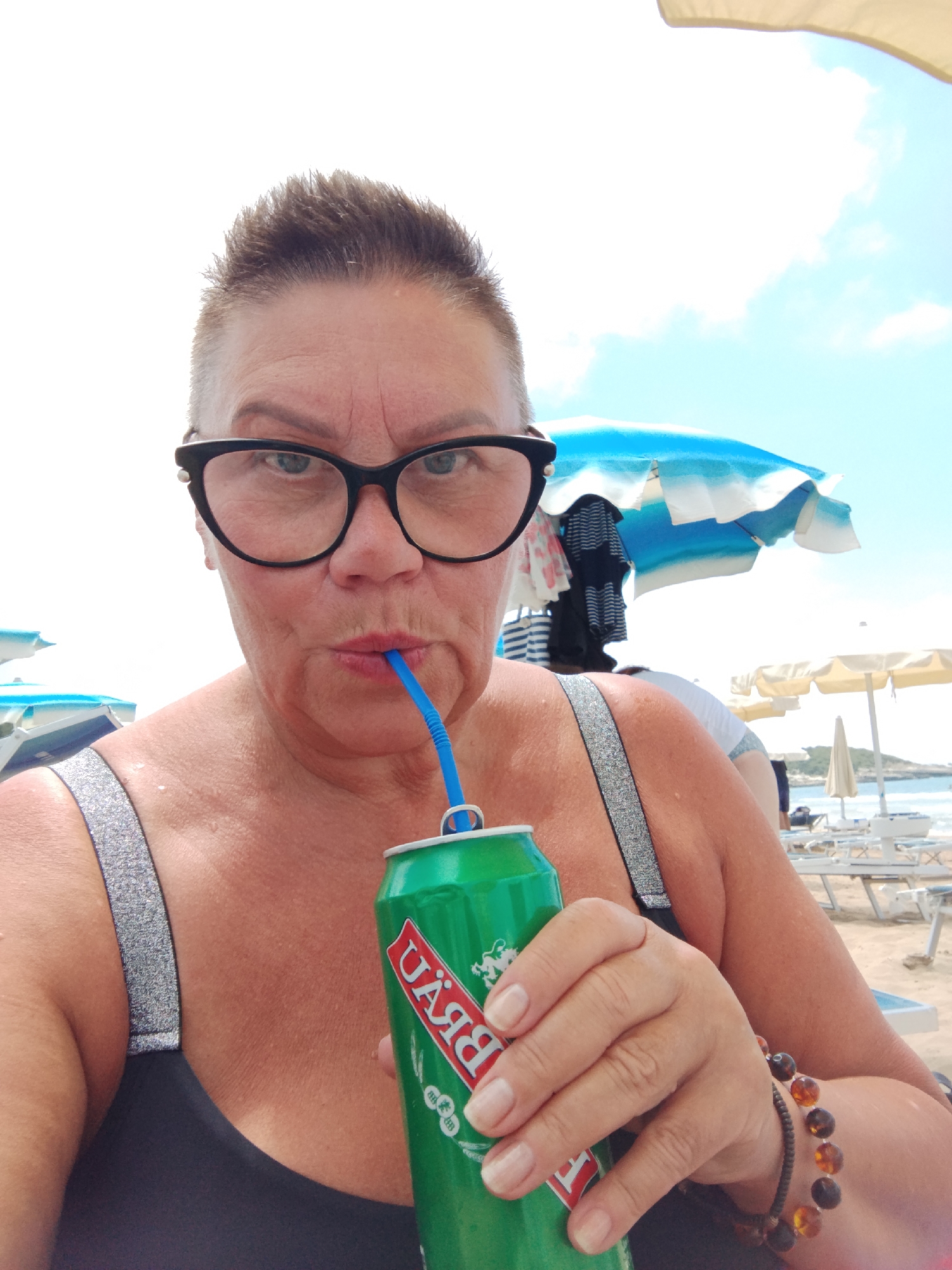 A woman with short, spiked hair and black-rimmed glasses is taking a selfie on a sunny beach in Peschici, Italy. She is wearing a black swimsuit with sparkly straps and is holding a green can with a blue straw to her lips. The can has red and white text on it, appearing to be a brand name. Her right hand, adorned with an amber beaded bracelet, is also holding the can.
The background shows a bright sky with scattered clouds. Beach umbrellas, some blue and white, and others yellow, are visible, along with lounge chairs, suggesting a typical beach day scene. The water and coastline can be seen in the distance. The overall mood is relaxed and summery. The lighting indicates it is daytime, likely late morning or early afternoon given the bright sunshine.