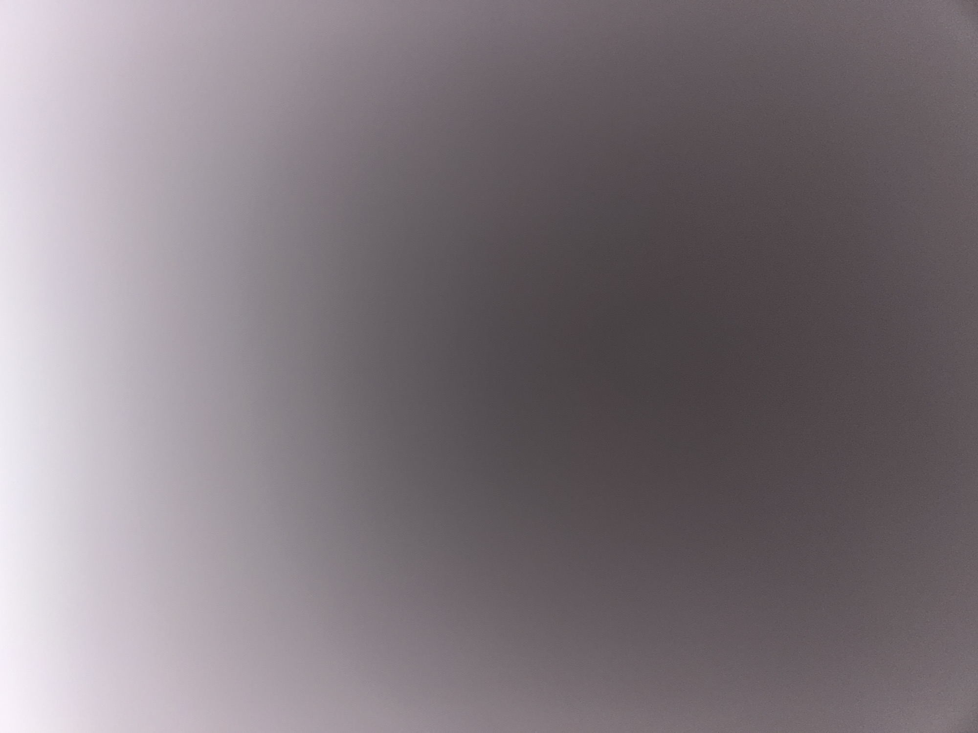 The image is a close-up shot of a textured surface with a gradient of colors, ranging from a light, almost white shade at the top to a darker gray at the bottom. The texture appears to be granular, with small, subtle bumps or irregularities visible throughout the surface. The lighting creates a soft, diffused effect, with no harsh shadows or bright highlights. There are no discernible objects, people, or any identifiable features within the frame, making the scene abstract and devoid of context.