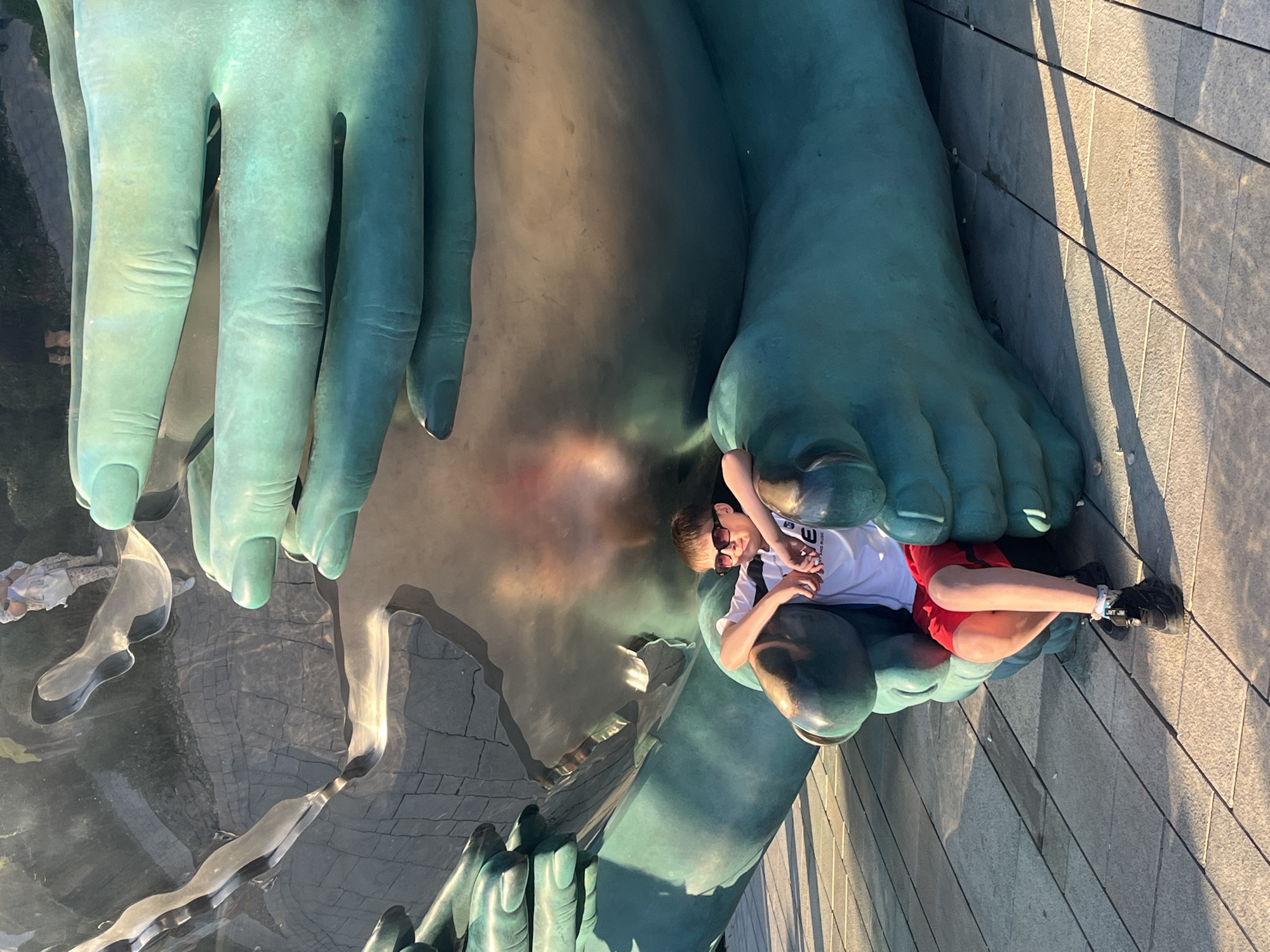 The image, taken in Krasnodar, Russia, features a young boy interacting with a colossal, green-patinated sculpture of human body parts, specifically a hand and a foot. The boy, appearing to be a pre-teen or teenager, has light brown hair, wears sunglasses, a white t-shirt, red shorts, and black sneakers. He is seated casually on the big toe and forefoot of the giant sculpture, resting his arms on its lower leg, suggesting a relaxed and playful engagement with the artwork. A small white bracelet is visible on his right ankle.
The sculpture itself is made of a green, metallic-looking material, likely oxidized bronze, indicating a public art installation. The immense scale of the hand and foot dominates the left and center of the frame. The sculpture is situated outdoors on what appears to be a highly reflective surface, possibly a shallow pool of water or polished metal, which mirrors distorted reflections of the boy, the sculpture, and another child in light clothing on the far left.
Bright sunlight illuminates the scene, casting strong shadows on the tiled or paved ground adjacent to the sculpture, suggesting a clear and sunny day, likely late morning or afternoon. The boy's t-shirt has a partially visible logo or text, including the letters "U" and "PURSUE". The overall atmosphere conveys a sense of leisure and interaction with large-scale public art.