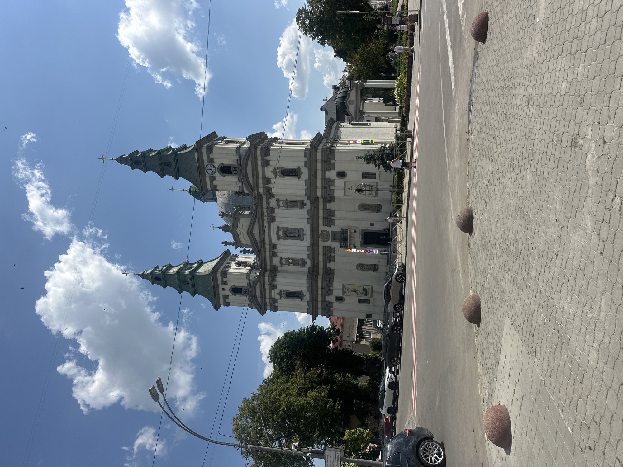 The image depicts the Church of the Immaculate Conception of the Blessed Virgin Mary in Ternopil, Ukraine, on a sunny day with scattered clouds. The church is a large, ornate building with two prominent green spires topped with crosses. It features a white facade with decorative moldings and several windows. A clock is visible on one of the spires.
In the foreground, a paved pedestrian area with interlocking stones is visible, with several large, rounded, reddish-brown bollards placed at intervals. A street with a pedestrian crossing runs across the image. Several cars are parked along the side of the street. Among the cars are a black Volkswagen Golf R, a white Toyota RAV4, a dark gray BMW X5, and a light beige BMW X3.
On the sidewalk in front of the church, several people are walking. A man in a white tank top and dark shorts is walking across the pedestrian crossing, and a few other individuals are seen in the distance near the church entrance and along the sidewalk. Trees and greenery surround the church and the adjacent area. Power lines and a street lamp are also visible in the sky. The overall scene captures a busy urban environment with a significant religious landmark.