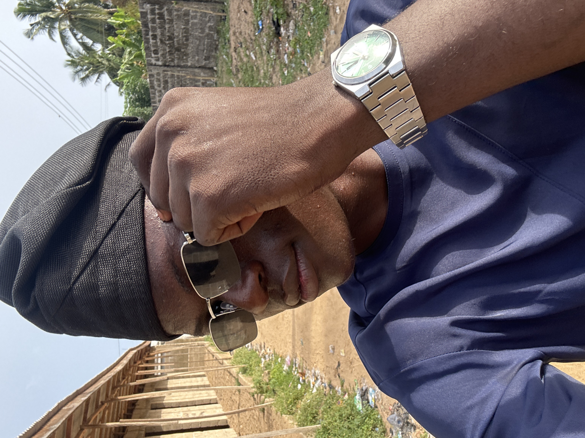The image shows a man with dark skin wearing a navy blue shirt and sunglasses. He has a black head covering and a silver wristwatch on his left wrist. His hand is raised near his forehead, possibly shielding his eyes from the sun. The background includes a partly constructed or deteriorated wooden structure, vegetation, and debris suggesting a somewhat unkempt environment. In the distance, there appear to be palm trees and utility poles, suggesting a tropical or coastal location. Without specific landmarks or additional context, confirming the precise location within Badagry, Nigeria, is not possible based solely on the image.